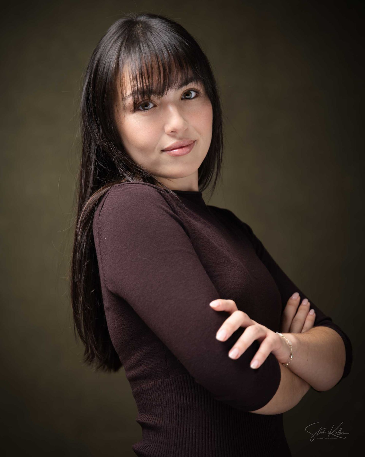Young woman with dark hair and bangs in a brown ribbed top, arms crossed, posing confidently against a dark studio background.