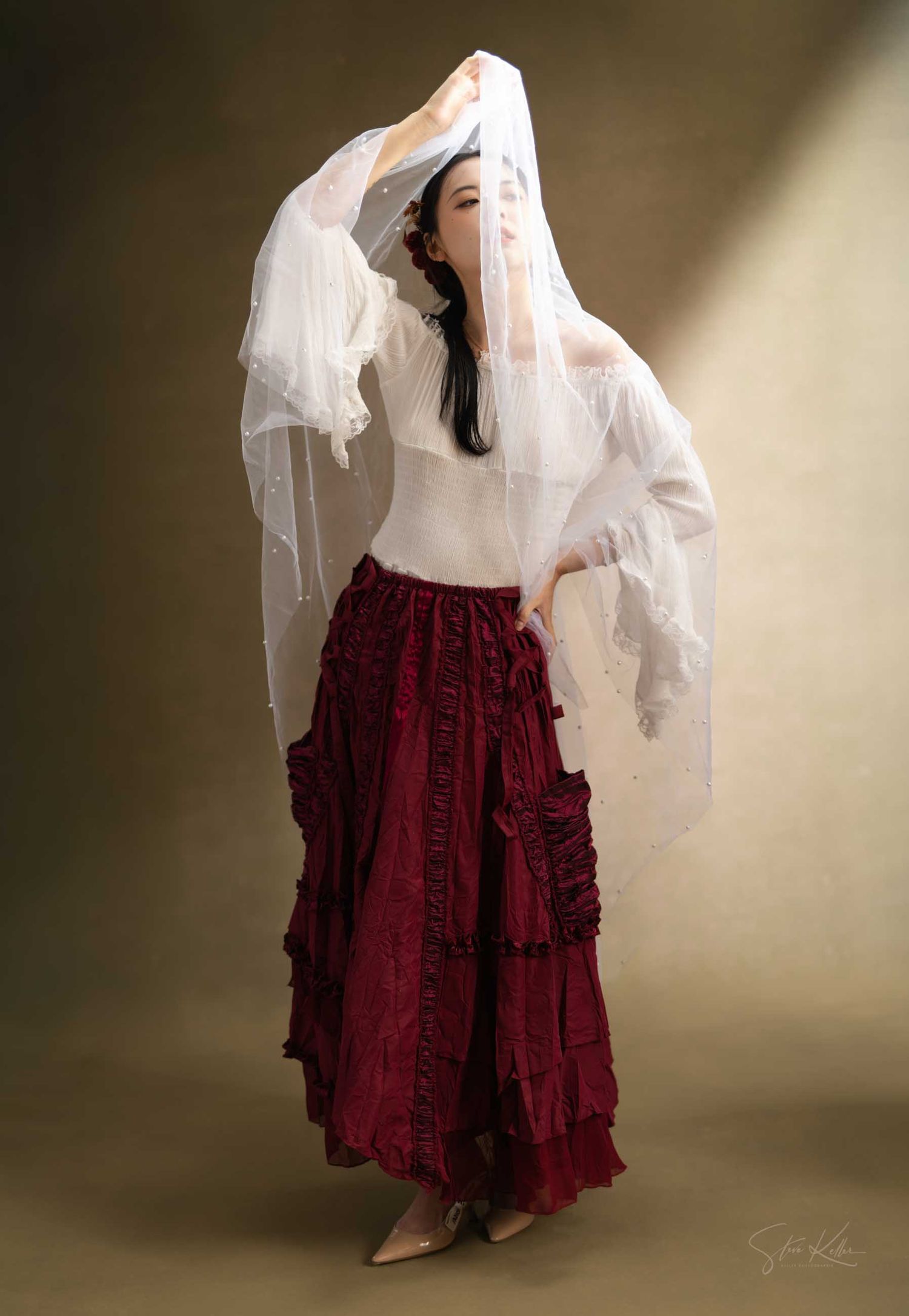 Figure in flowing white top and burgundy ruffled skirt poses dramatically with sheer fabric against moody backdrop.