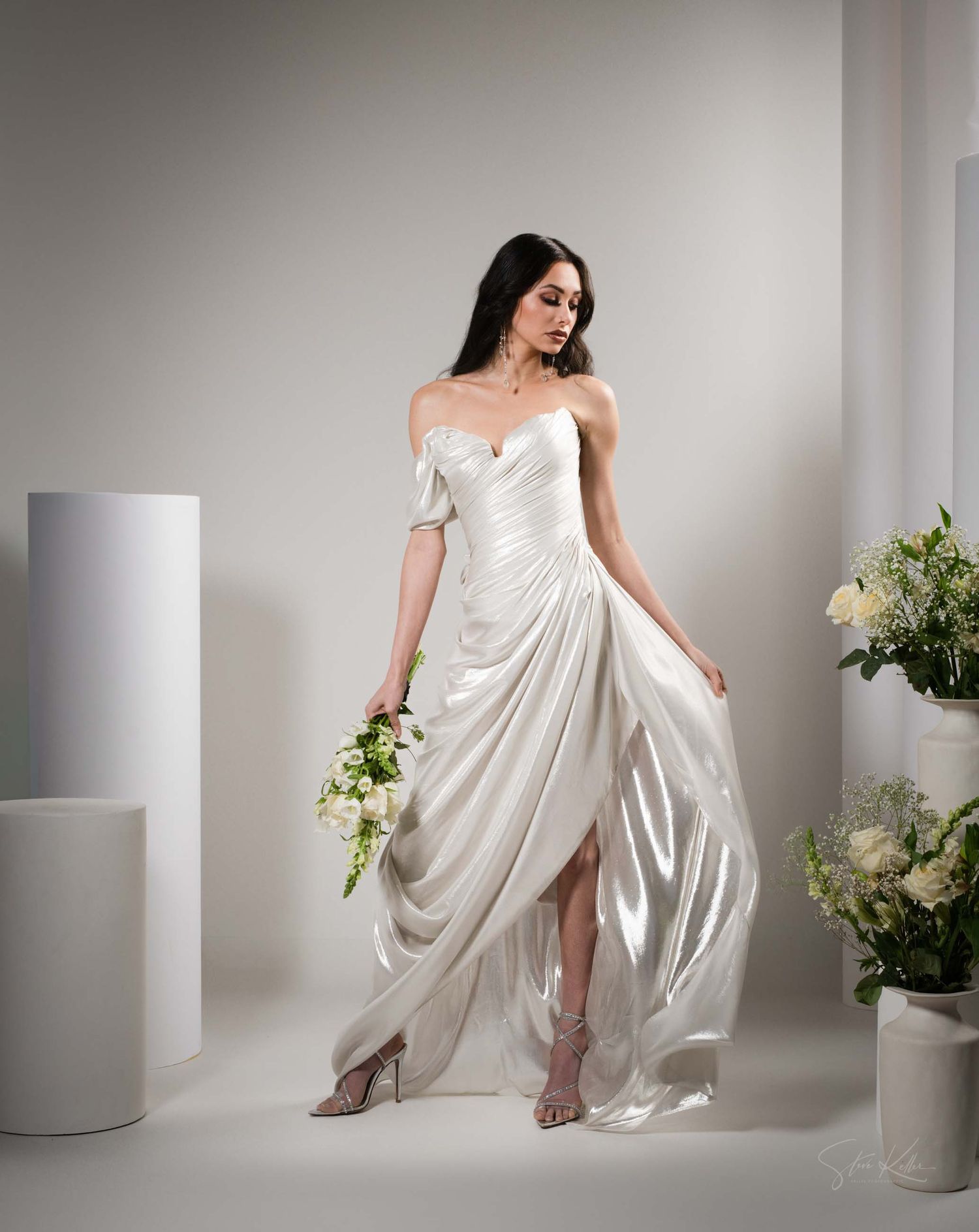 A flowing white satin dress creates an elegant silhouette against a minimalist studio backdrop with delicate floral accents.