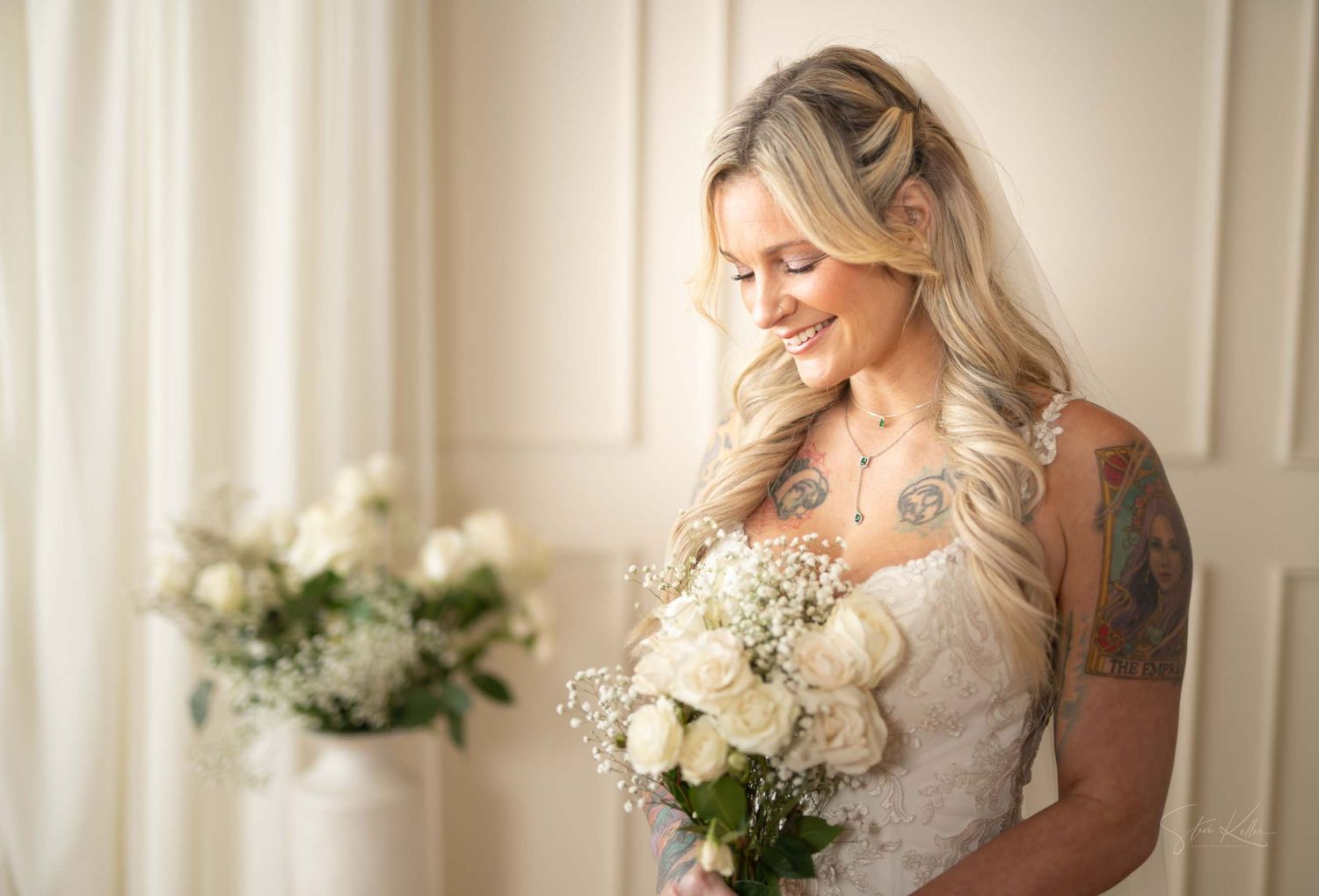 A bride with flowing blonde hair and tattoos gazes down at her white floral bouquet.