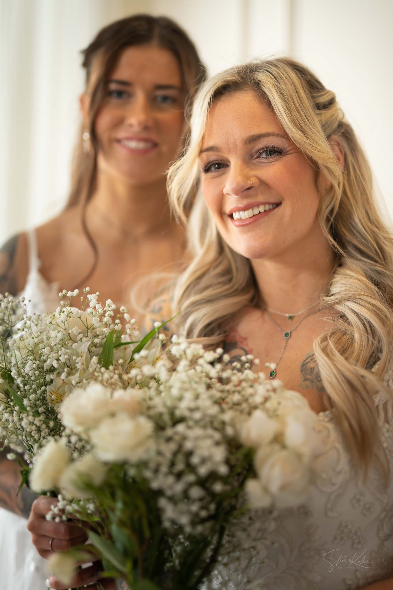 Two women in wedding attire smile while holding a bouquet of white roses and baby's breath flowers.
