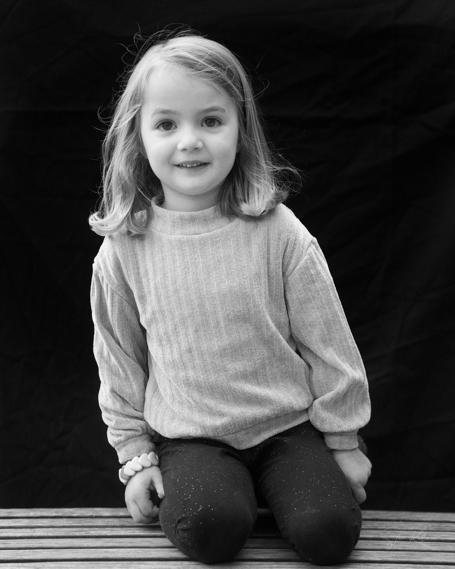 A child wearing a light gray sweater sits on a wooden bench in a black and white photograph.