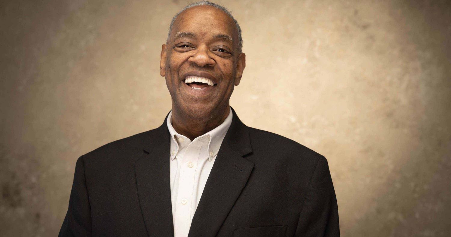 Smiling older man in a black blazer and white open-collar shirt laughs warmly against a textured beige background.