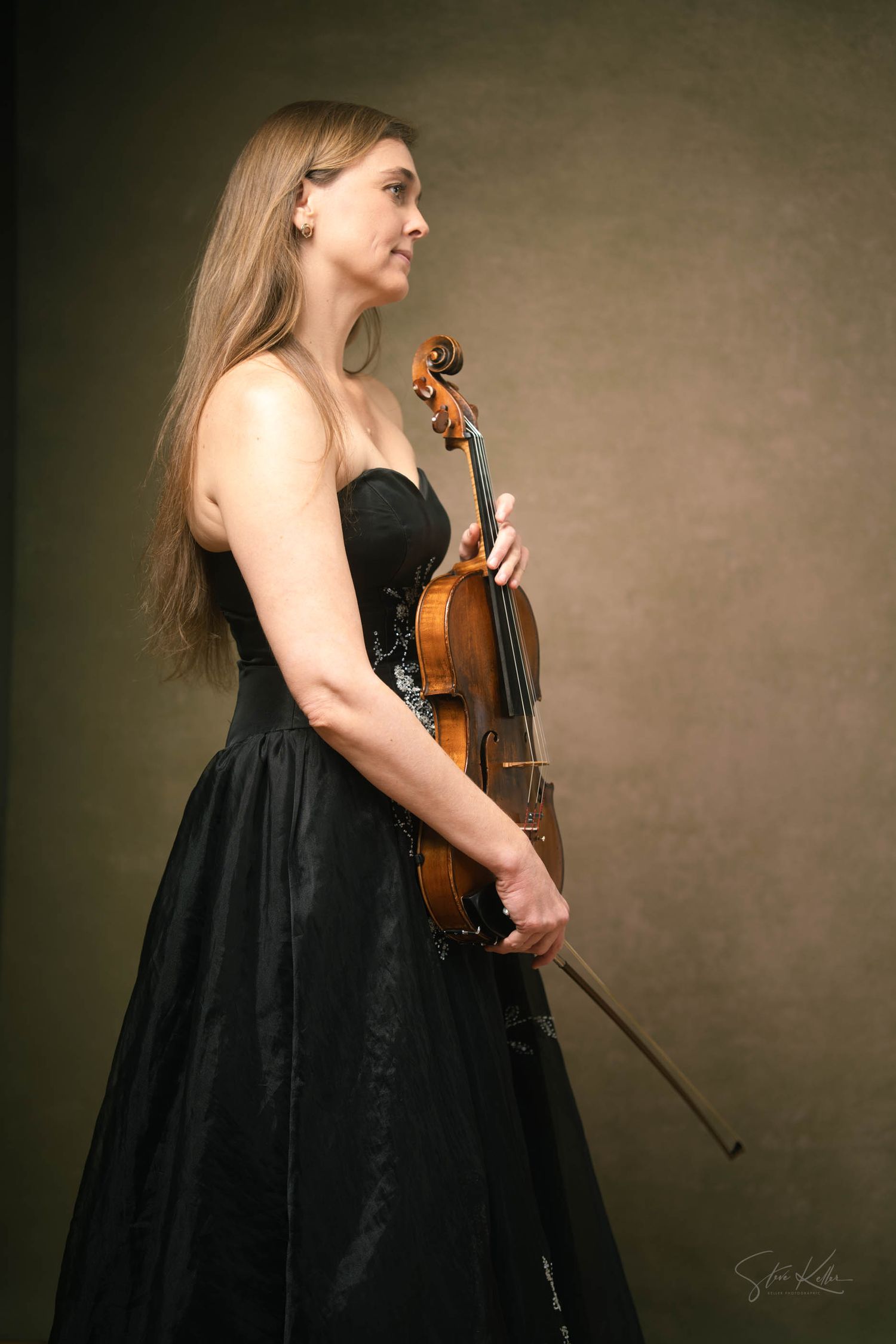 Classical musician in elegant black dress poses with violin against dark backdrop in artistic portrait series.