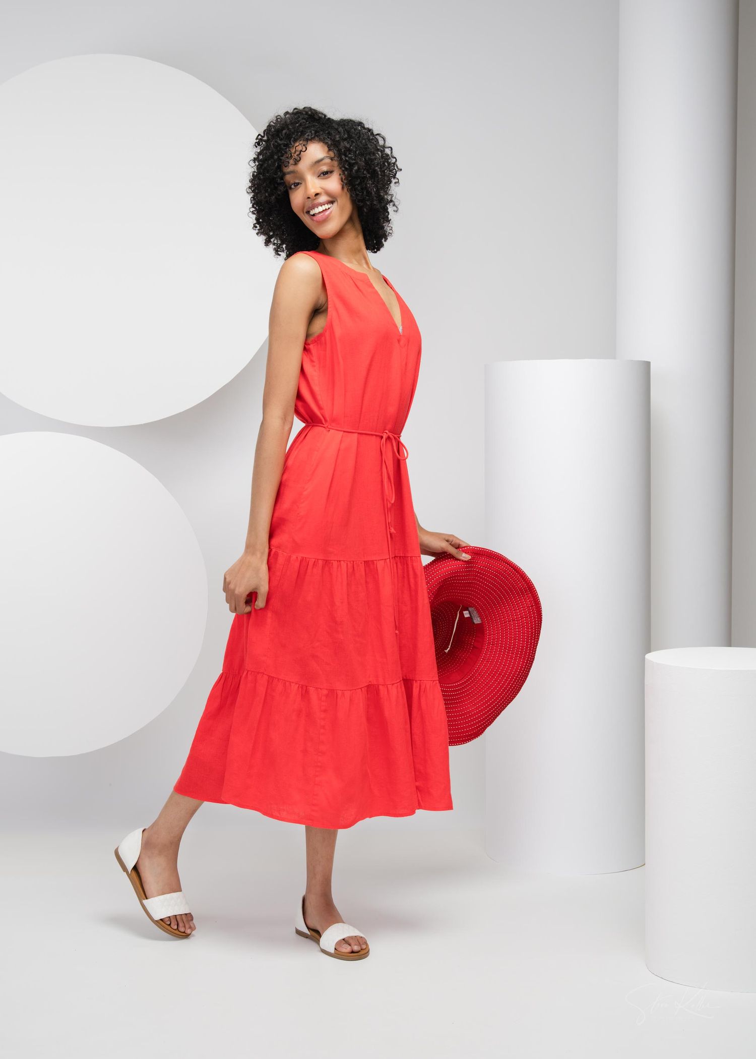 Person in vibrant red tiered maxi dress with white sandals poses against white geometric backdrop.