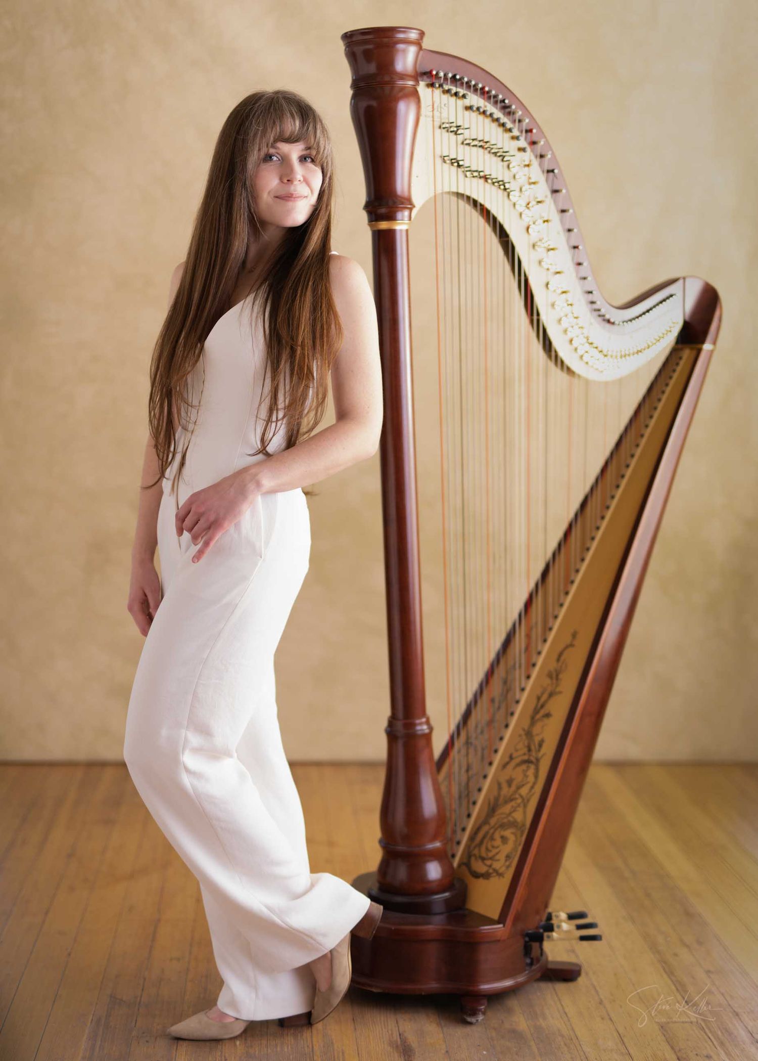 A female harpist in white attire stands elegantly beside a tall wooden concert harp on a wooden floor.