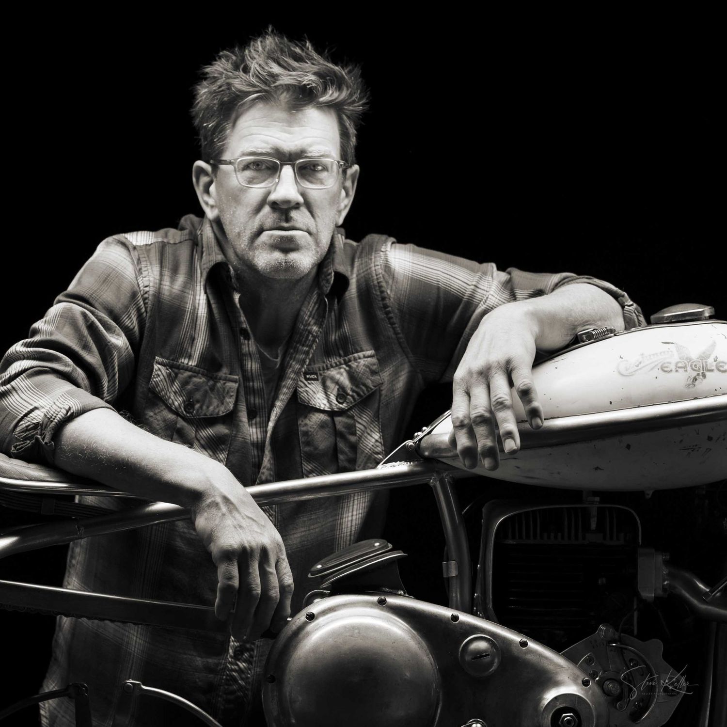Black and white portrait of a middle-aged man leaning on a vintage motorcycle, wearing a denim jacket.