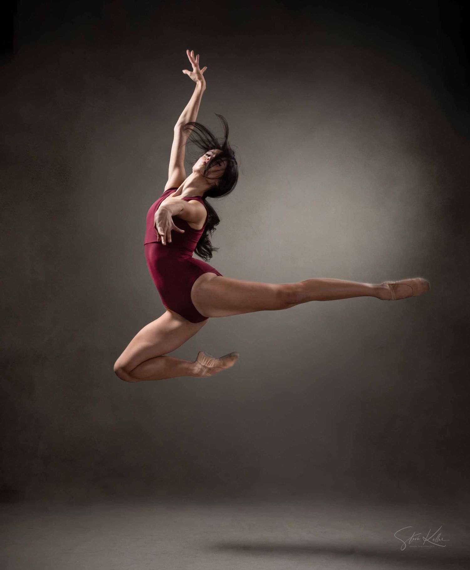 Dancer in burgundy leotard executes dynamic mid-air leap with extended limbs in dark studio setting.