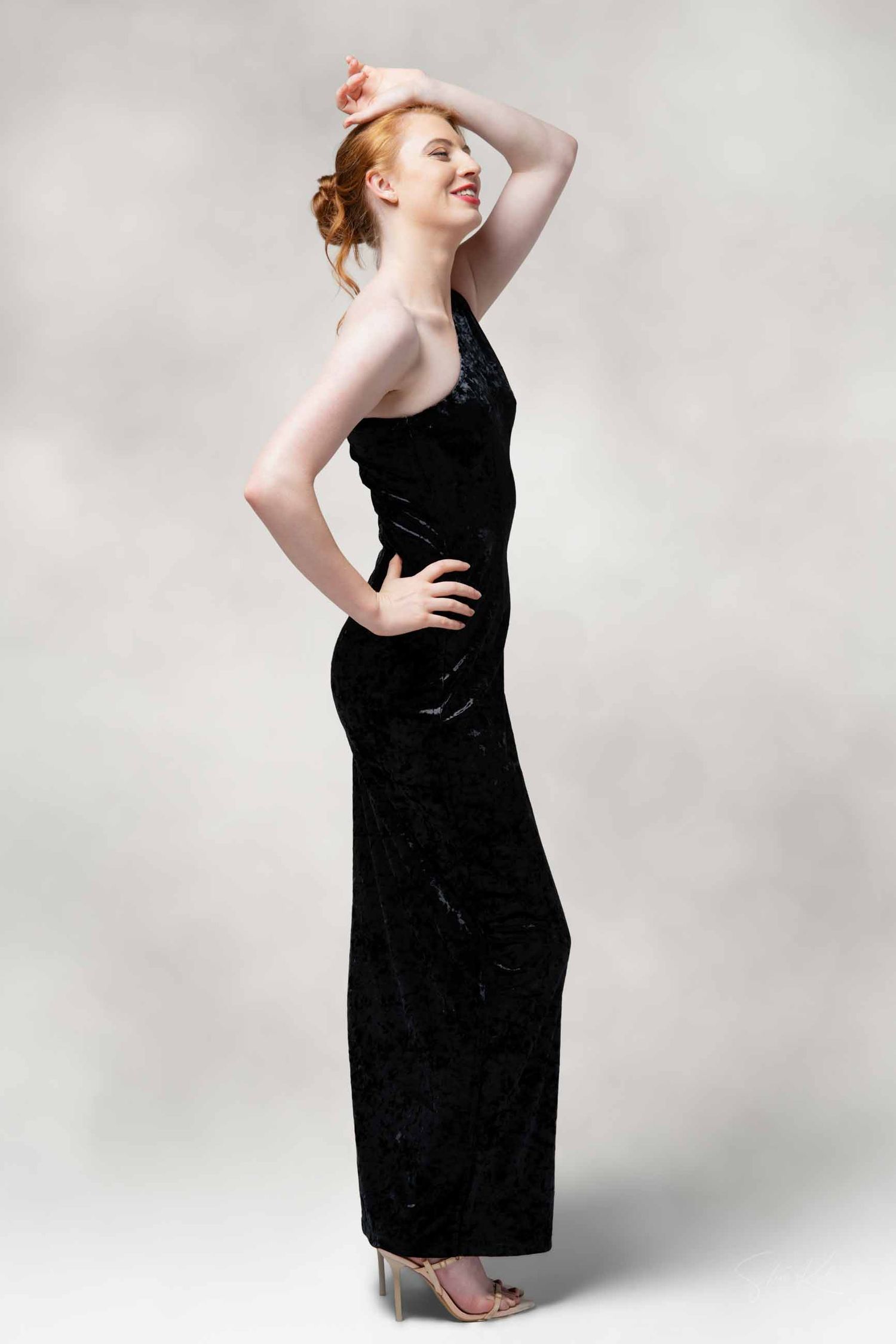 Model wearing black velvet evening dress poses dramatically against white background in studio setting.