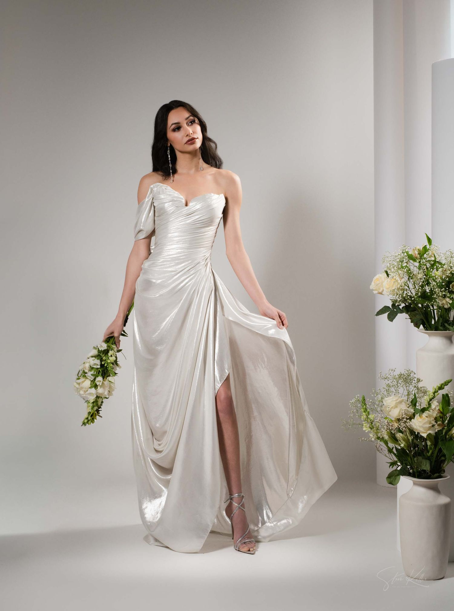 Elegant white flowing dress with draped fabric detail photographed in studio setting with potted white flowers.