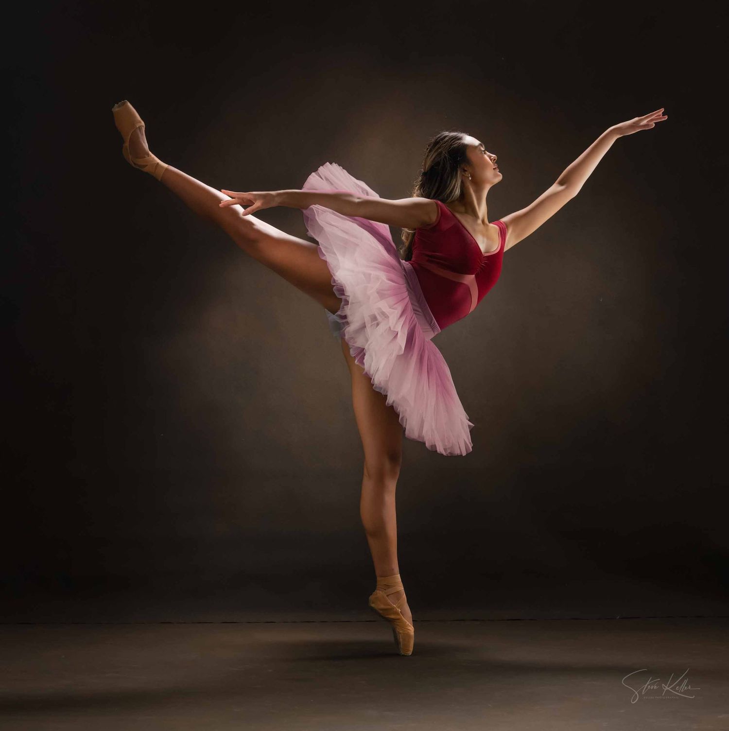 Ballerina in pink tutu performs arabesque pose on pointe against dark dramatic backdrop.