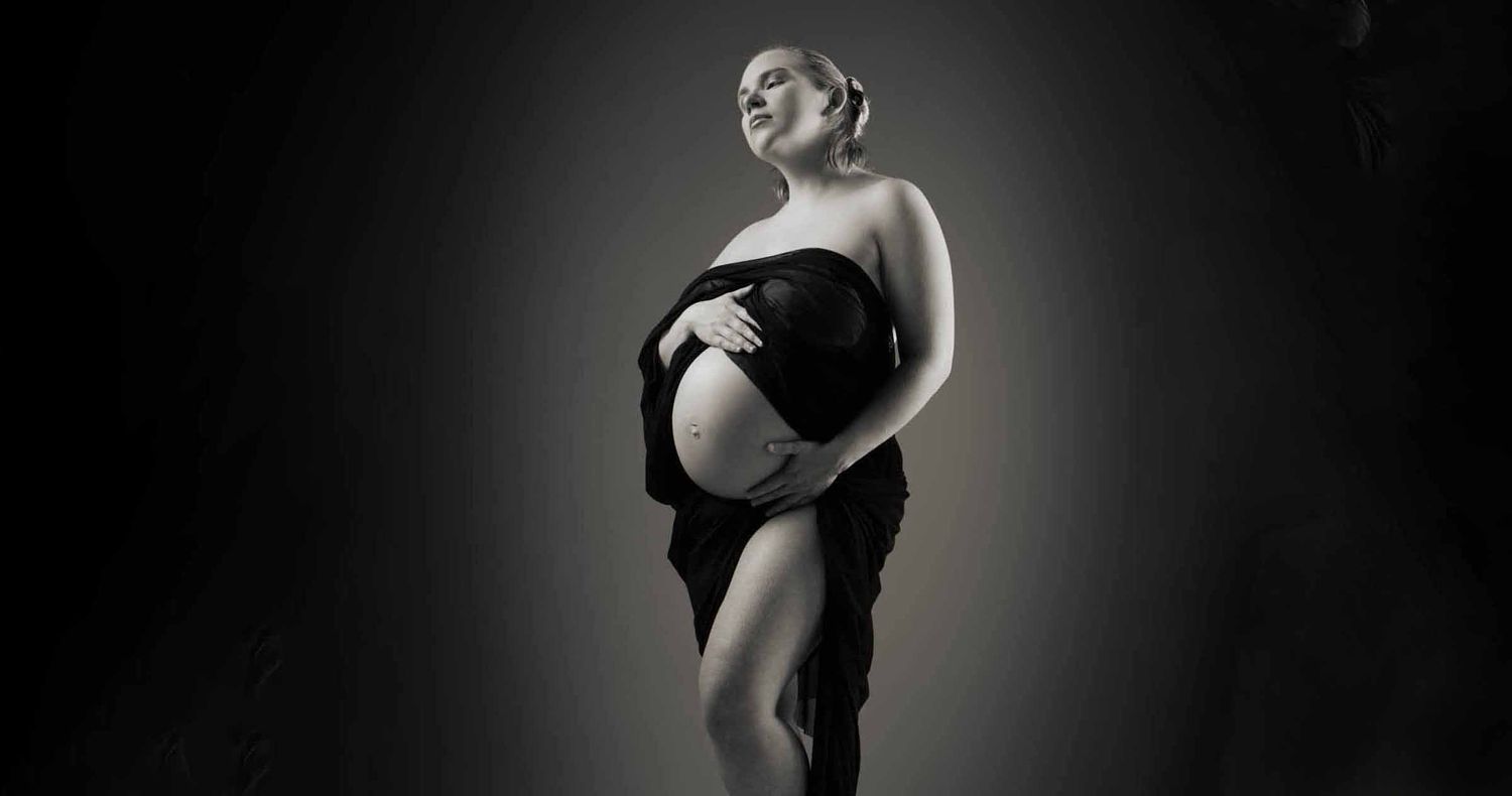 Pregnant woman in a black draped fabric cradles her bare belly in a dramatic black and white maternity portrait.