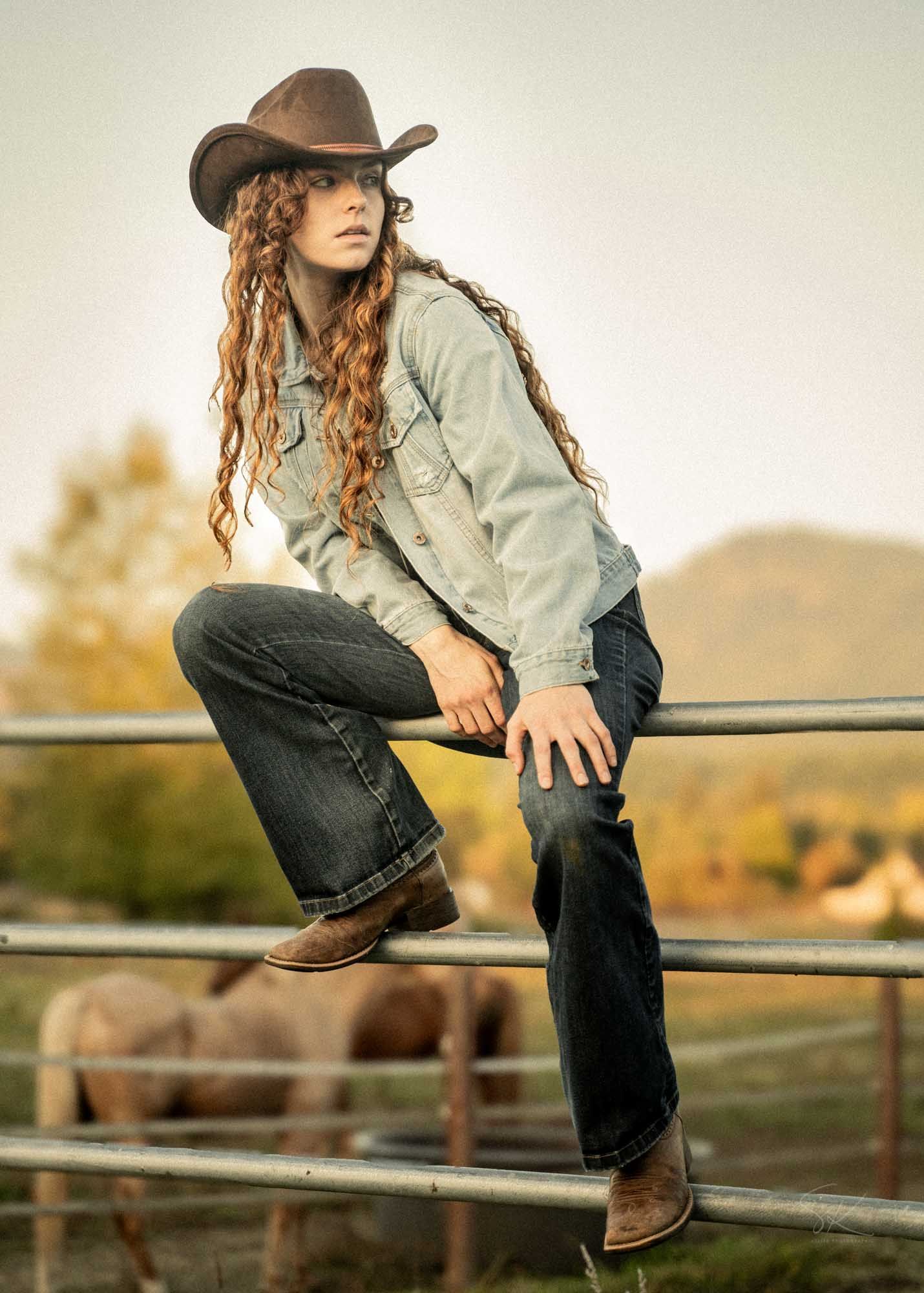 Young woman in brown cowboy hat and denim jacket sitting on metal ranch fence with livestock in background.