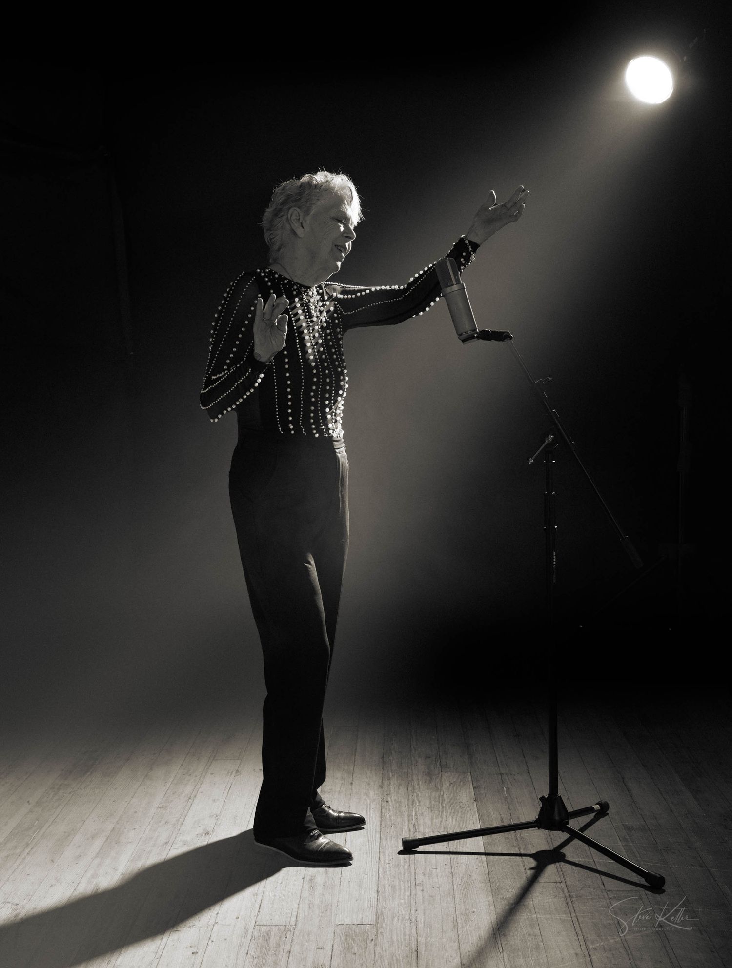 A dramatic black and white performance photo with a single spotlight illuminating a performer on a wooden stage.
