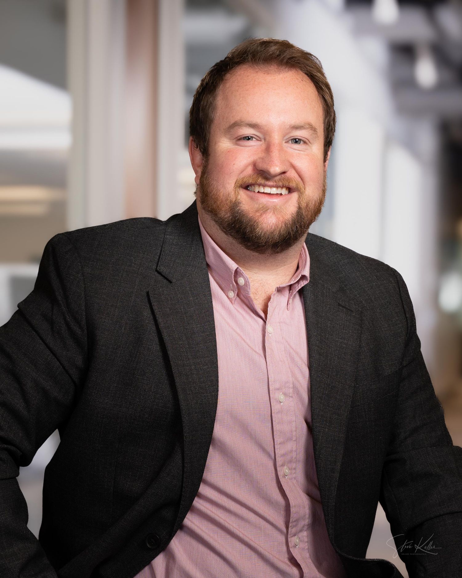 Professional headshot of person in black suit jacket and pink button-down shirt against light background.