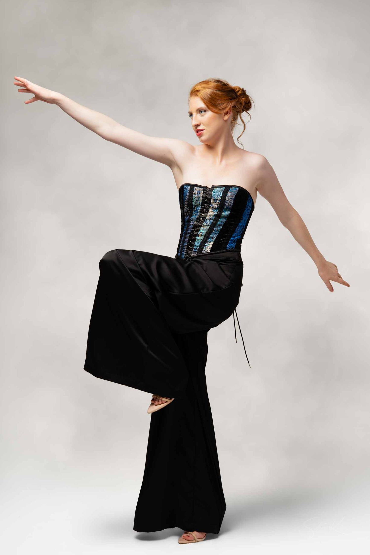 A dancer in a black dress with blue corset detail performs a graceful movement sequence against a light background.