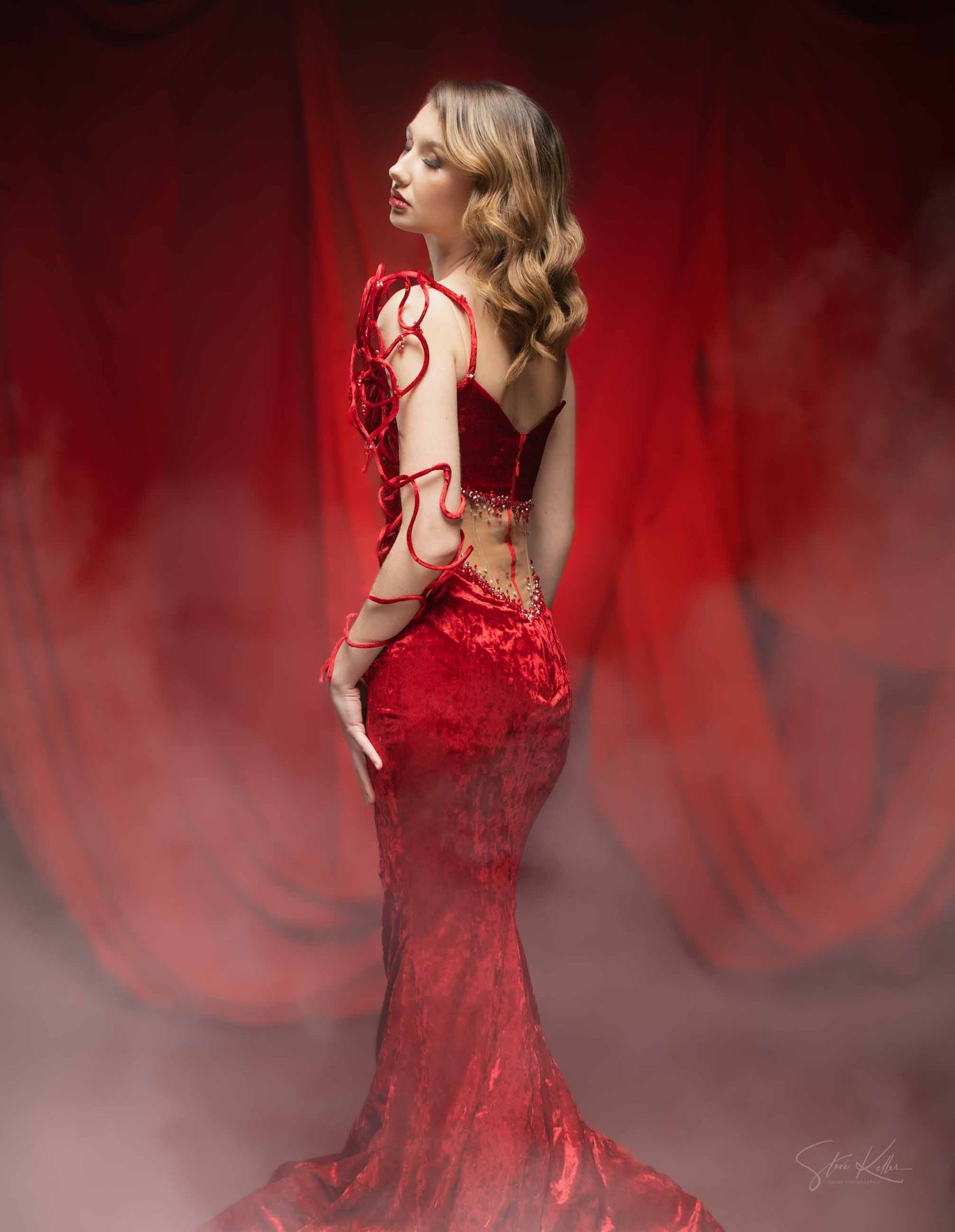 A model in a flowing red evening gown poses elegantly against a dramatic red and gray misty background.