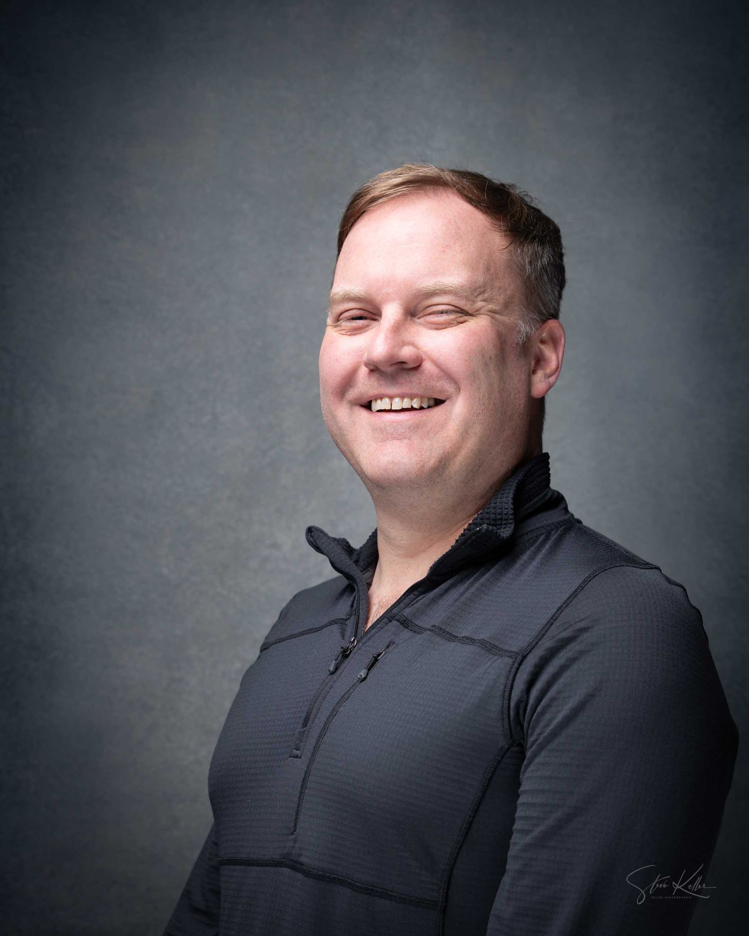 Smiling middle-aged man in a dark polo shirt poses confidently against a gray studio background.