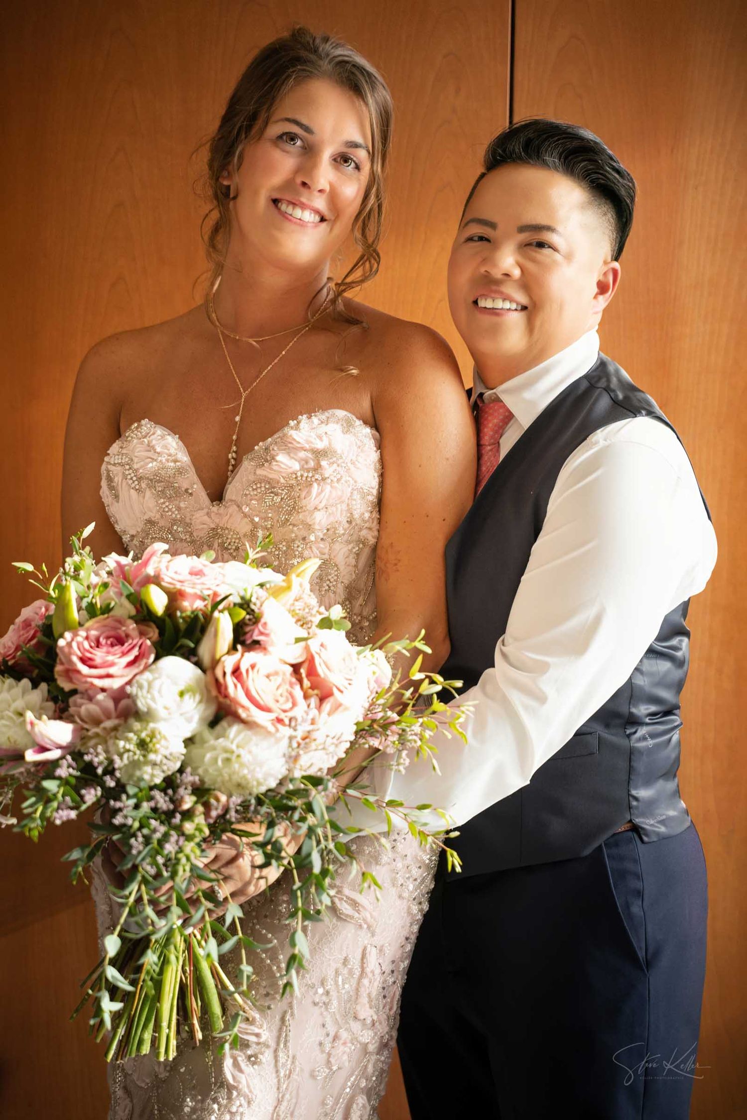 Two brides share a joyful moment indoors, one in lace gown with bouquet, other in navy vest and pink tie.