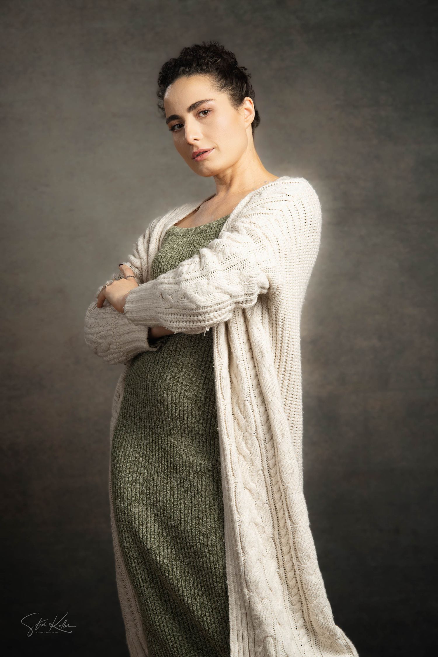 Maternity fashion portrait in olive dress and cream cable knit cardigan against dark backdrop.