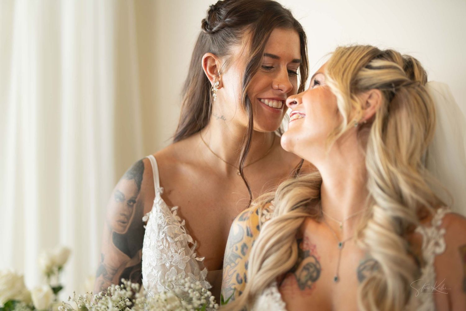 Two people share a tender moment in a light-filled room wearing elegant wedding attire.