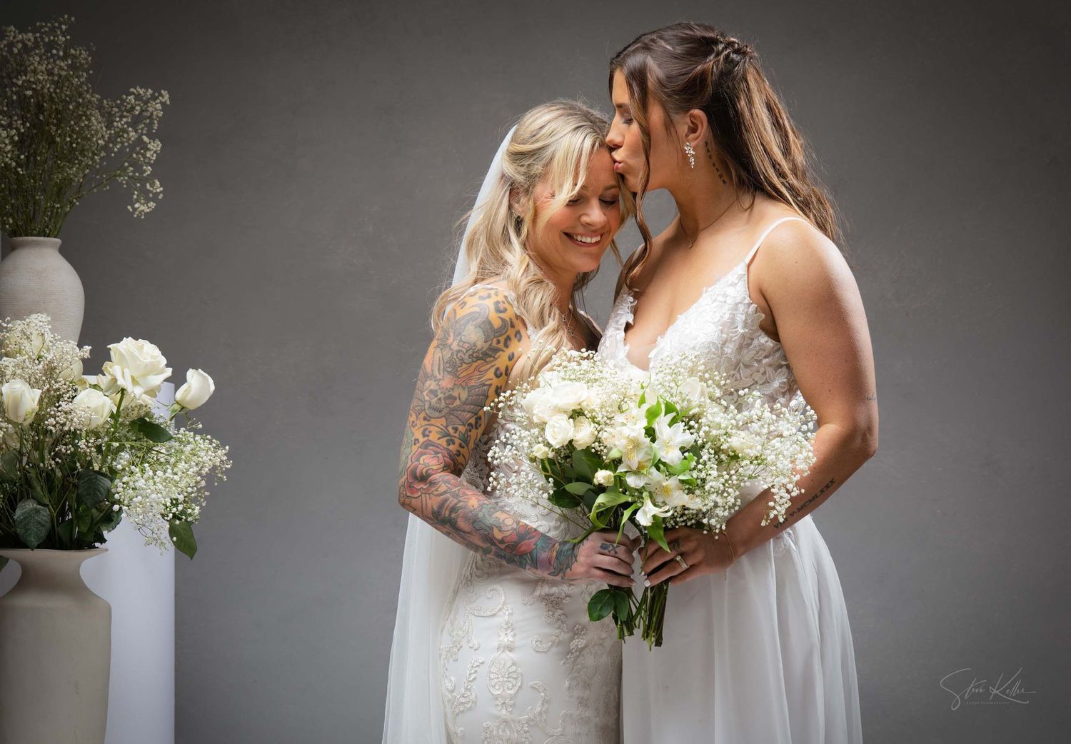 Two brides share an intimate moment at their wedding ceremony with white floral bouquets against a gray backdrop.