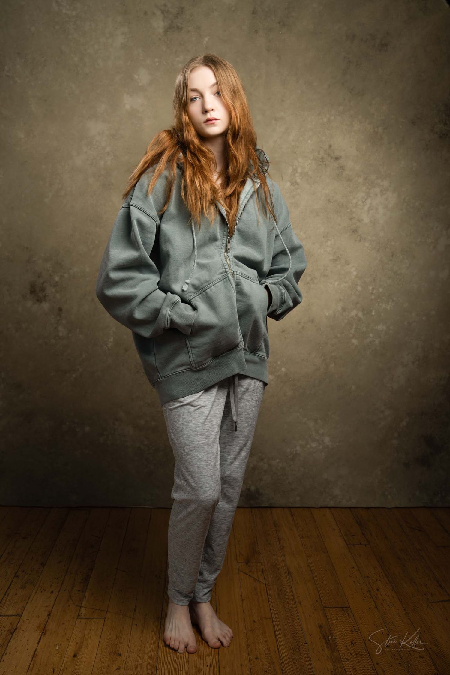Figure in baggy gray sweatsuit poses barefoot on wooden floor against textured wall in moody lighting.