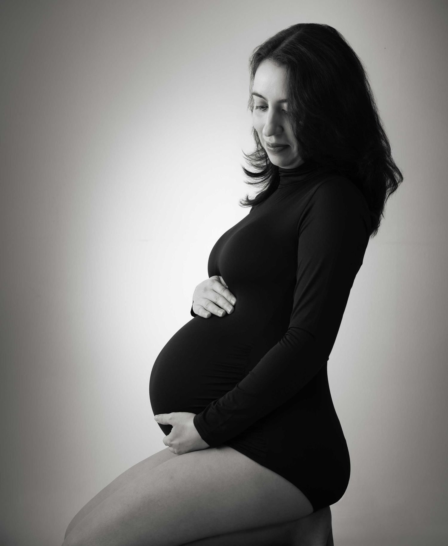 Black and white maternity photo of a pregnant woman in a black bodysuit cradling her baby bump in a studio.