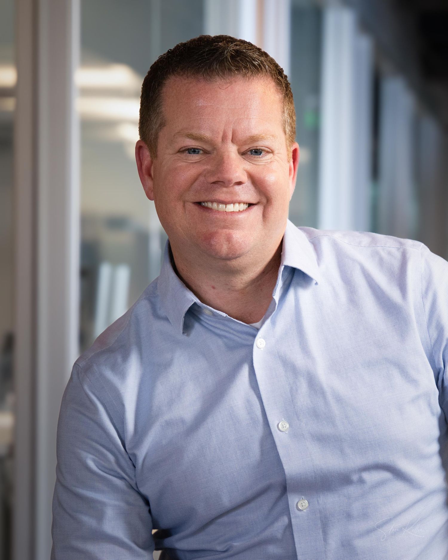 Professional headshot portrait of someone in a light blue button-down shirt against a blurred office background.