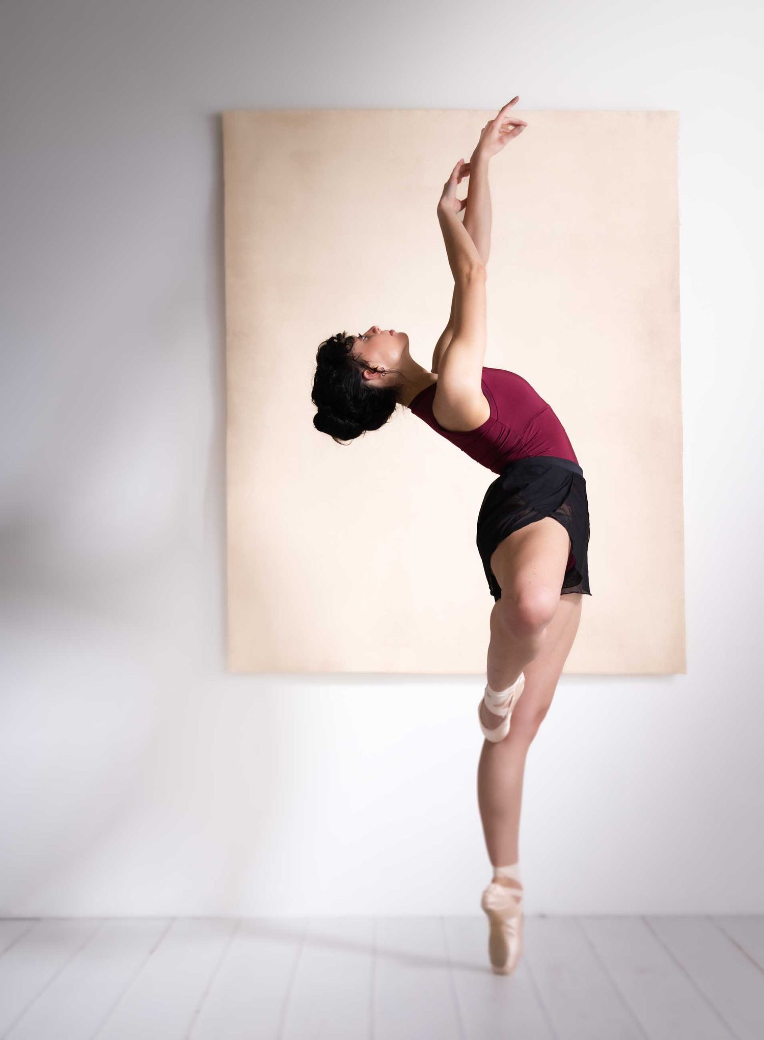 A ballet dancer in pointe shoes performs an elegant arabesque in a studio, wearing a red top and black skirt.
