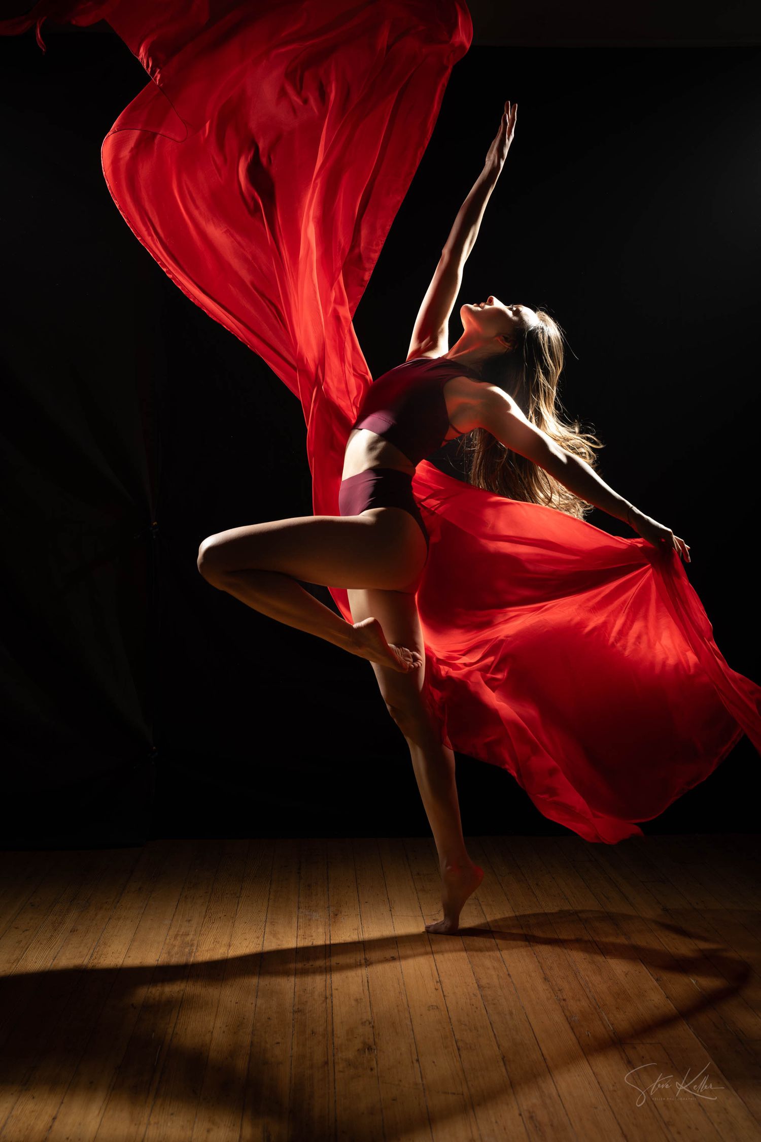 A dancer in red fabric performs an elegant leap on a wooden floor, with flowing crimson cloth creating dramatic movement.