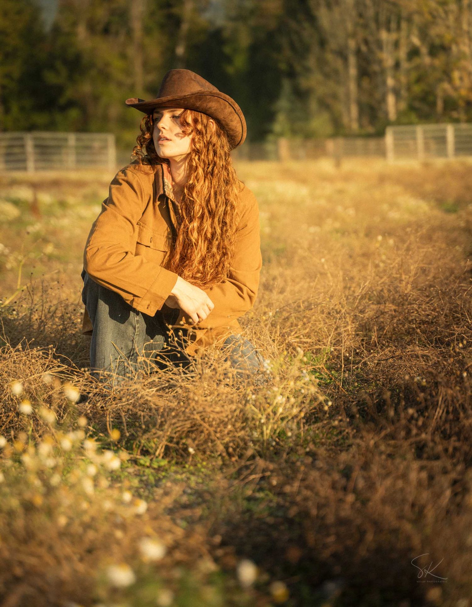 A young woman with long curly red hair wearing a brown cowboy hat and jacket sits in a golden field.