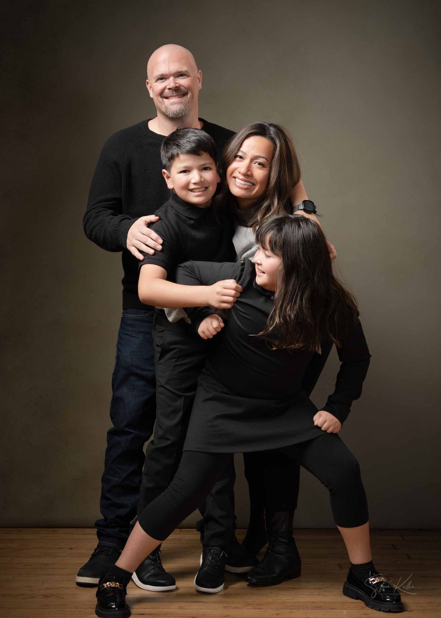 A family shares joyful embraces and laughter during a casual photo session against a dark backdrop.