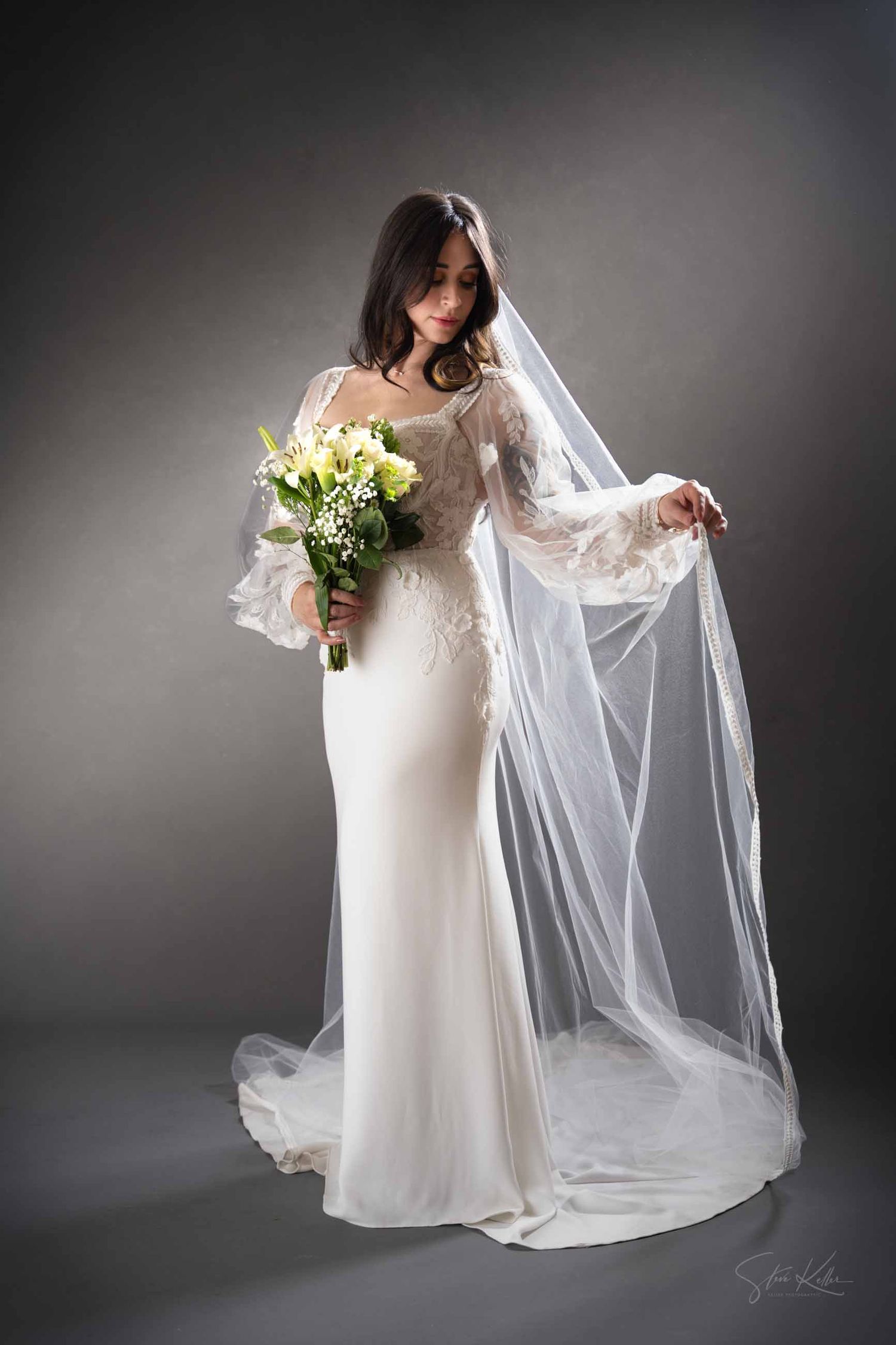 A bride in a flowing white gown holds a bouquet while gracefully posing with her long veil against a dark background.