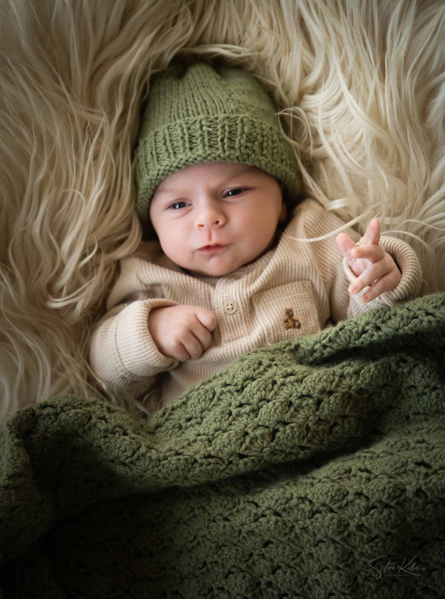 A baby wearing a knit beanie and sweater lies wrapped in cozy blankets against a soft furry background.