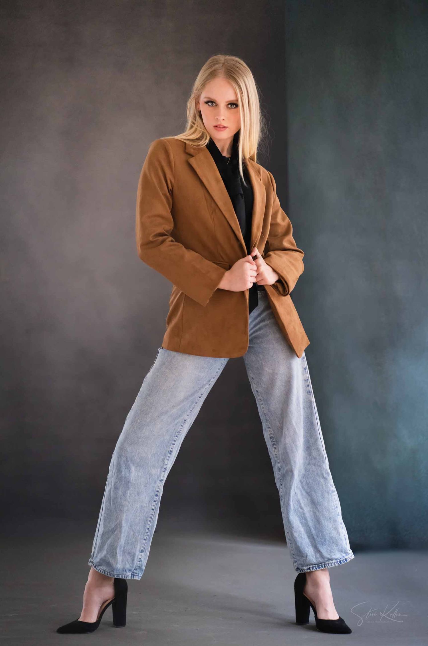 Person in brown blazer and light wash jeans poses dynamically against moody backdrop in fashion photoshoot series.