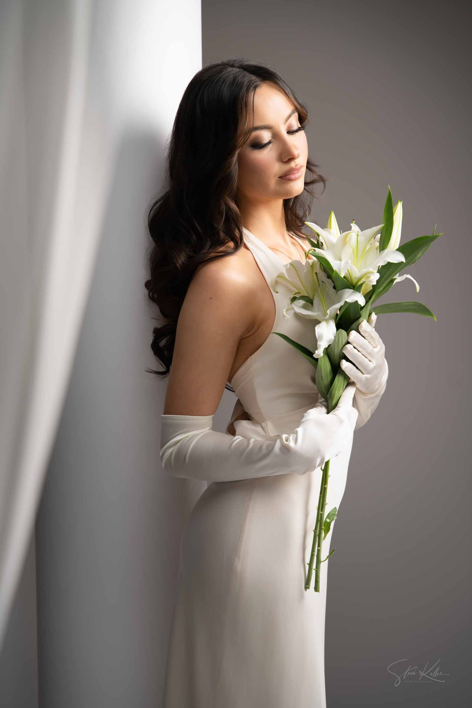 Elegant woman in white dress and long gloves holding white lilies in soft window light.