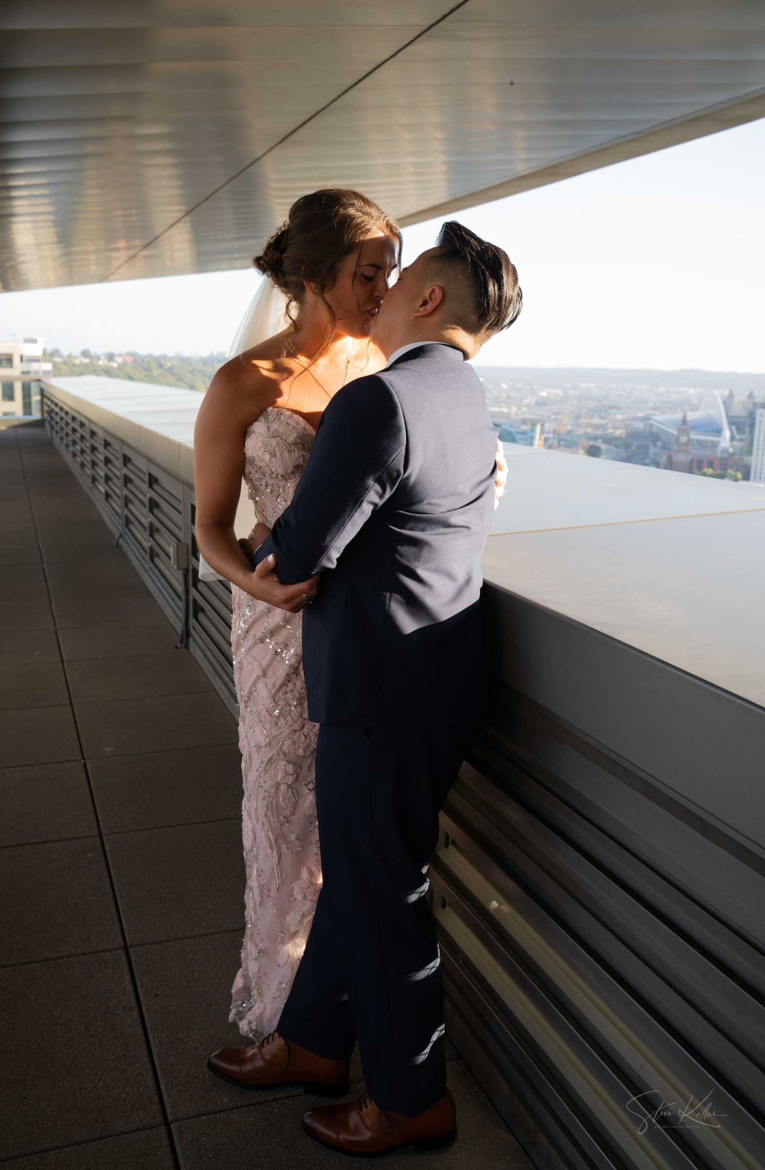 Couple sharing a romantic kiss on a rooftop terrace, bride in lace gown with veil, groom in navy suit.