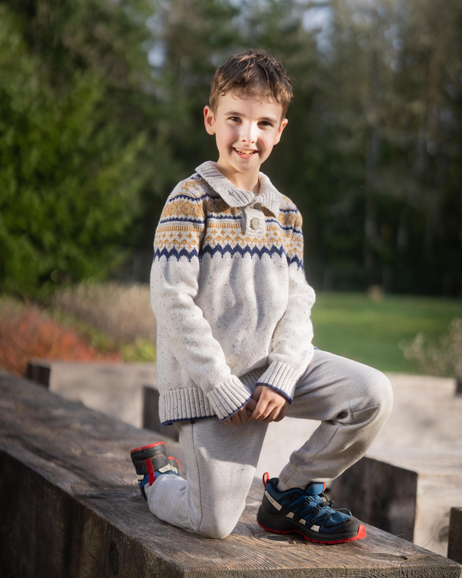 A young person in a patterned sweater sits casually on a stone wall outdoors in a park setting.