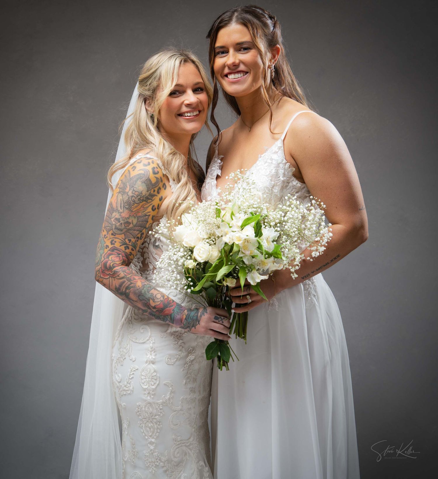 Two brides in white gowns hold bouquets and embrace during their wedding photoshoot.