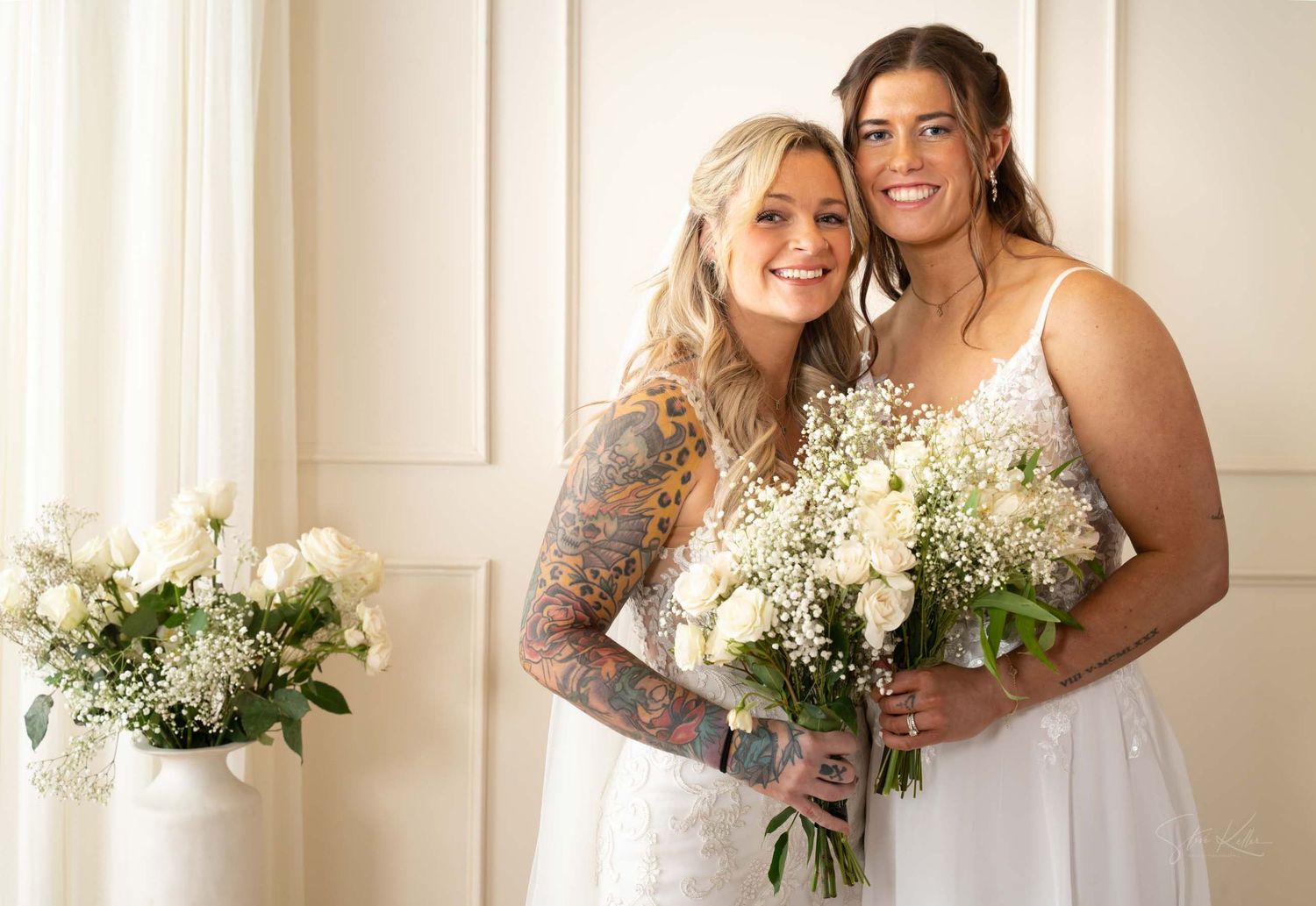 Two brides in white wedding dresses hold matching bouquets of baby's breath flowers while posing together indoors.