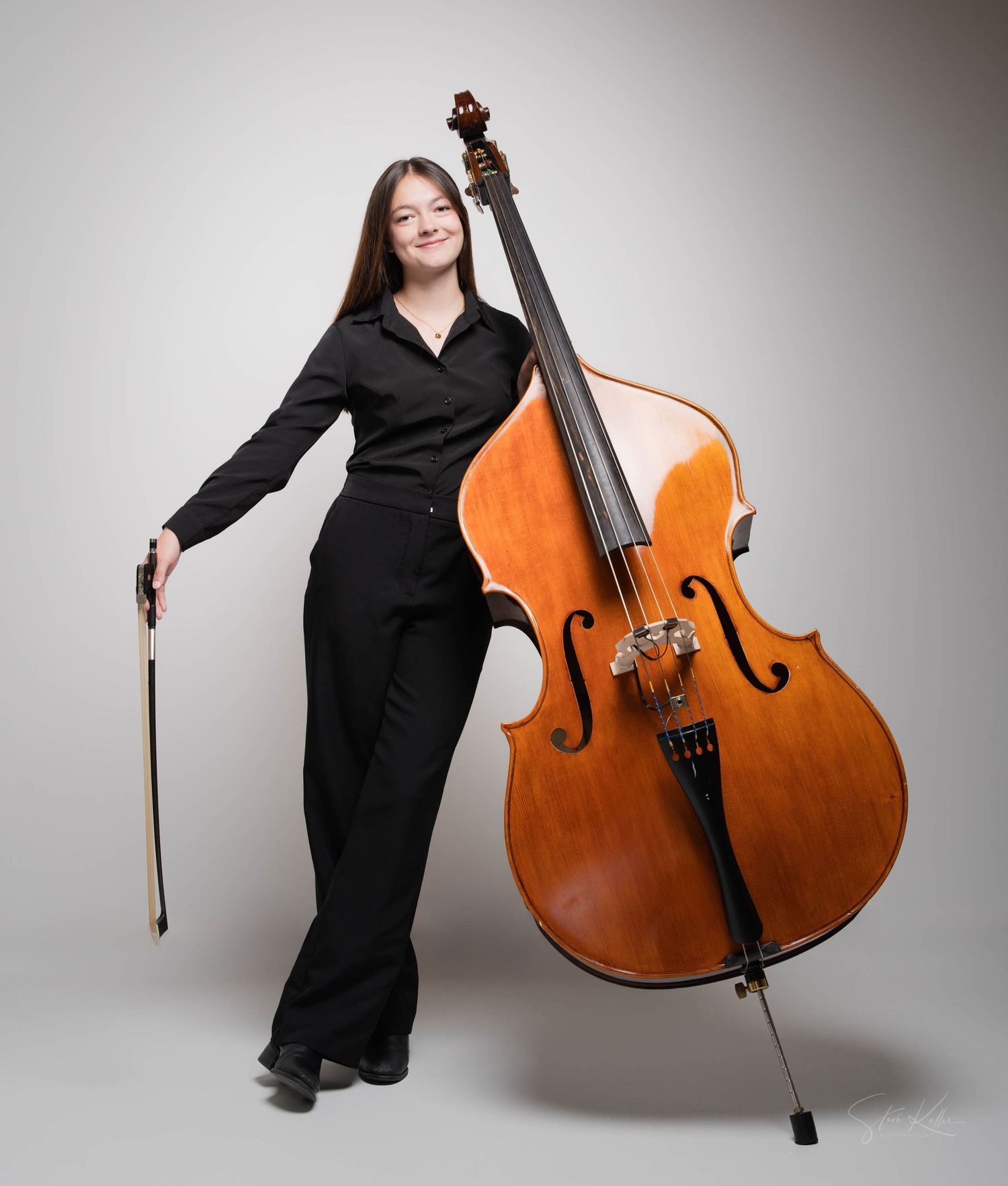 Musician in black formal attire poses elegantly with a large double bass instrument against a light gray studio backdrop.