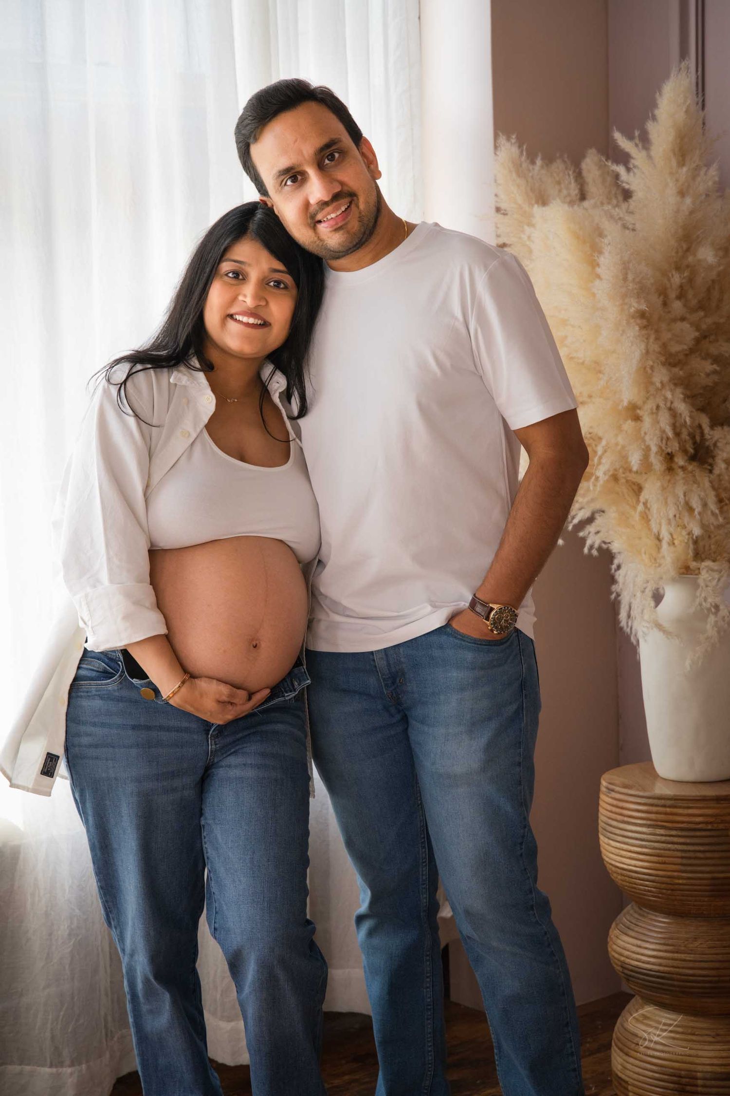 Happy couple posing for maternity photo, woman in white top showing baby bump, man in white shirt, both wearing jeans.