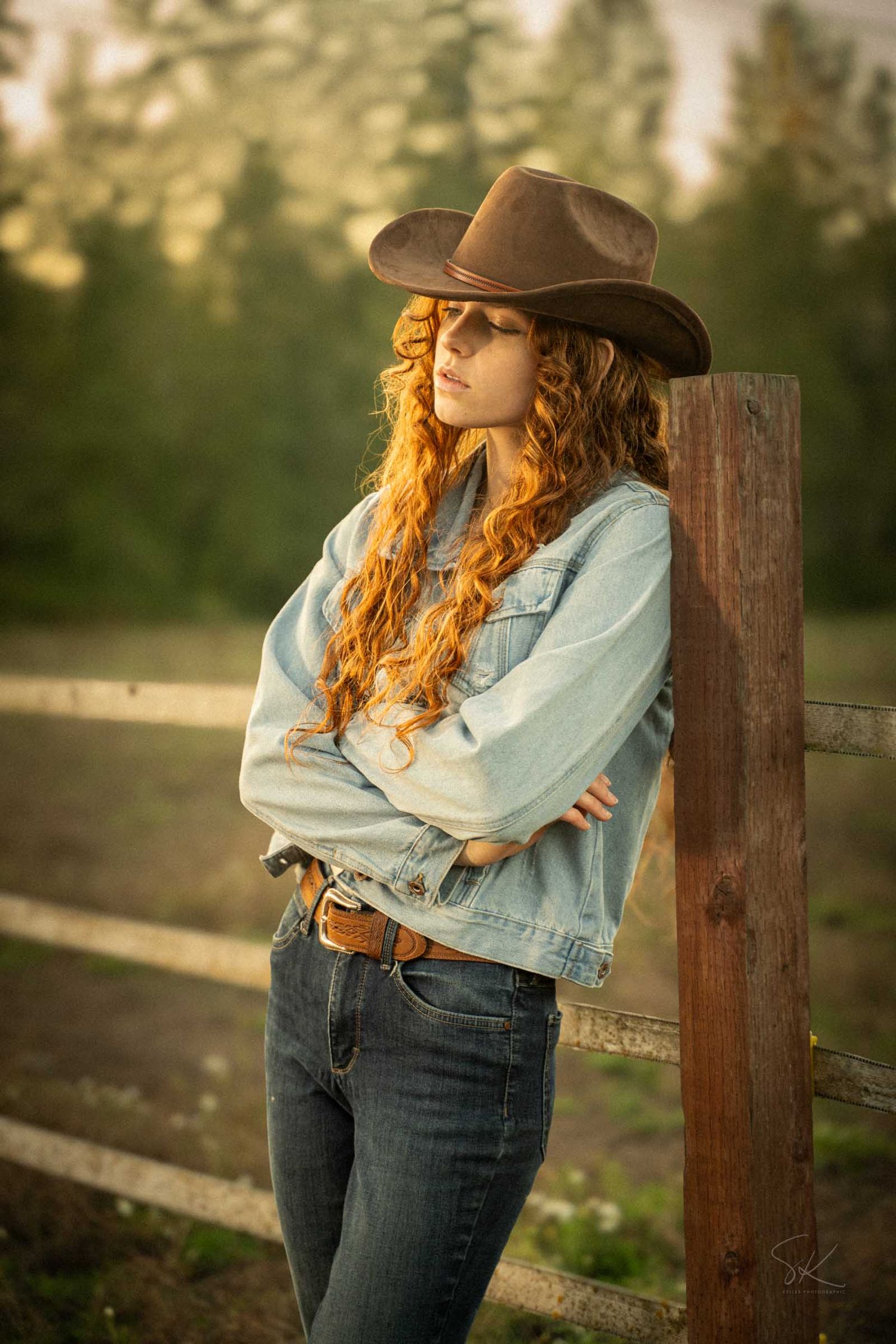 A young redhead woman in a cowboy hat, denim jacket, and jeans leans against a wooden fence post outdoors.