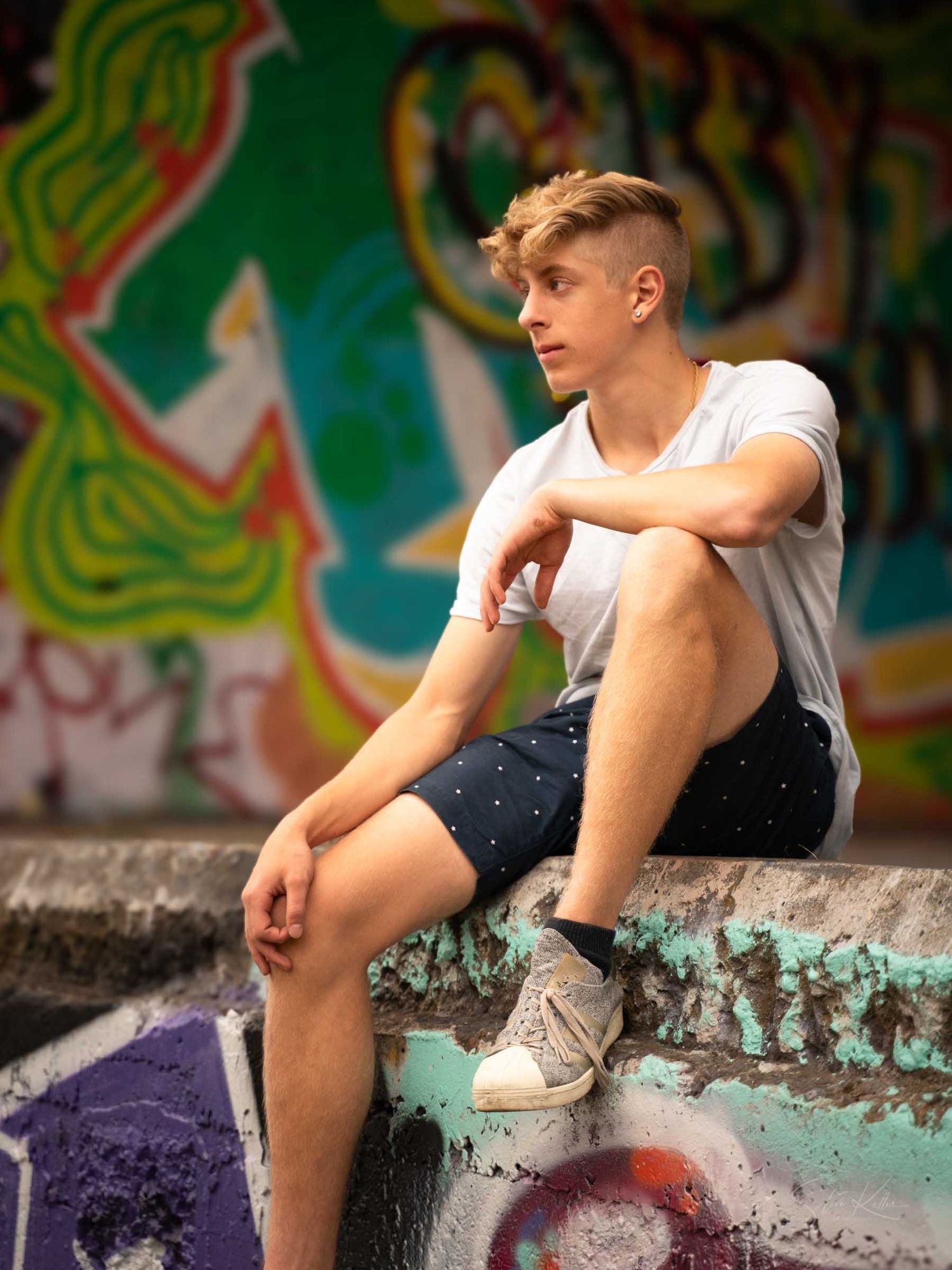 Young man in white t-shirt and dark shorts sitting on graffiti wall, gazing thoughtfully in urban setting.