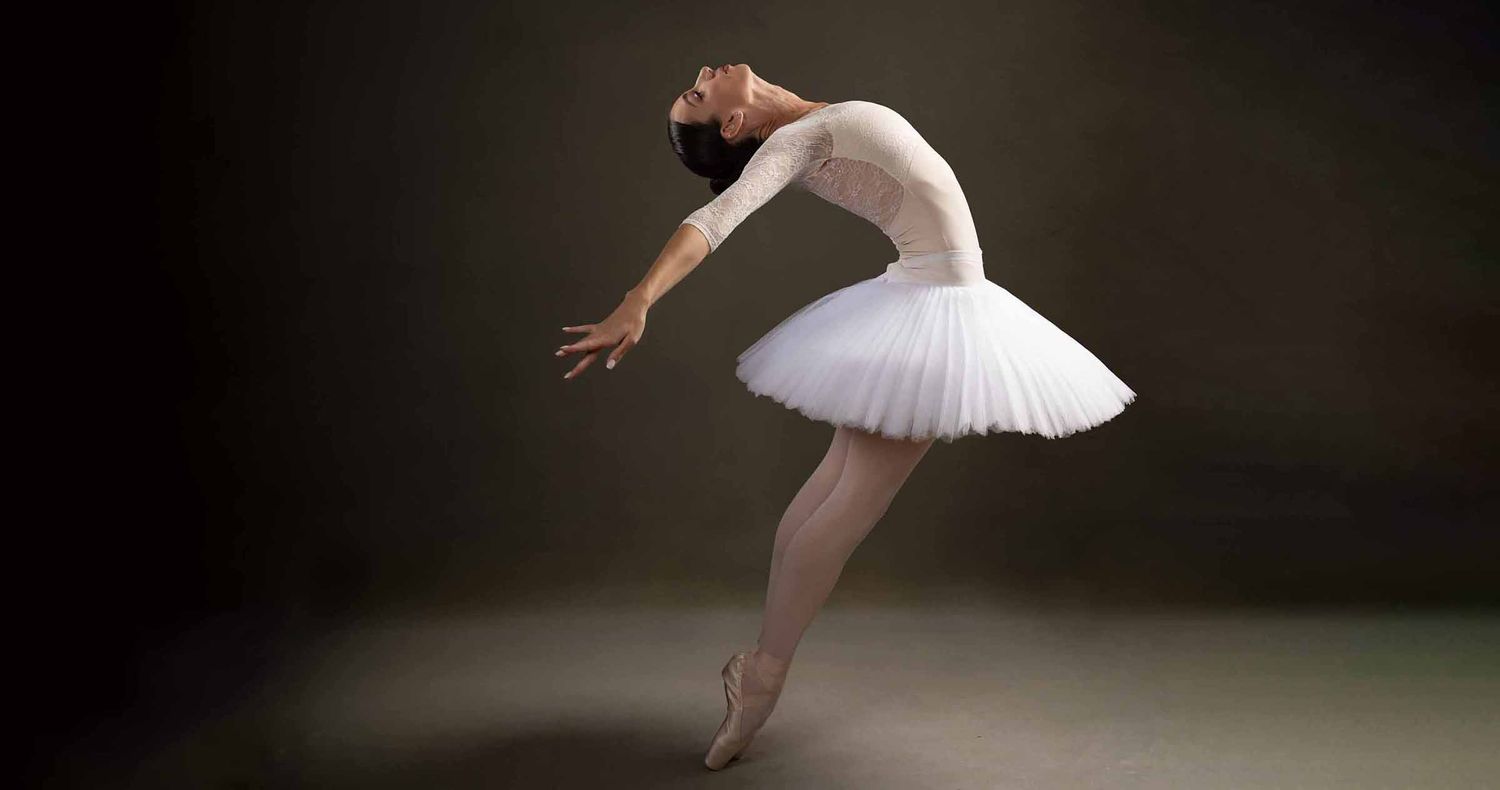 Ballerina in a white tutu arches her back gracefully en pointe against a dark, moody studio backdrop.