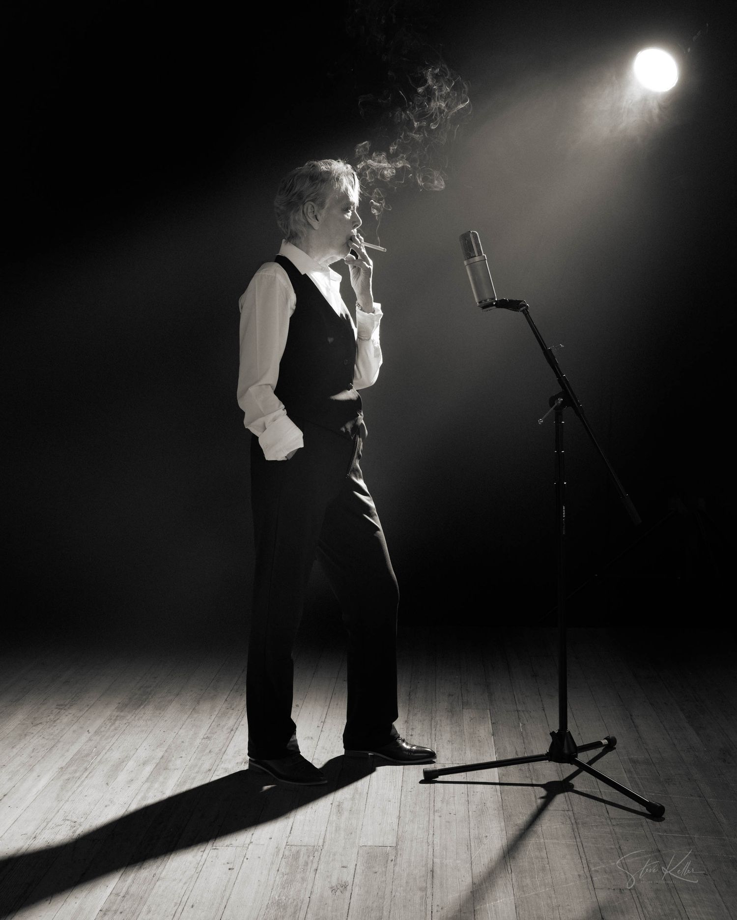 A performer in formal attire stands on a wooden stage under dramatic spotlight next to a microphone stand.
