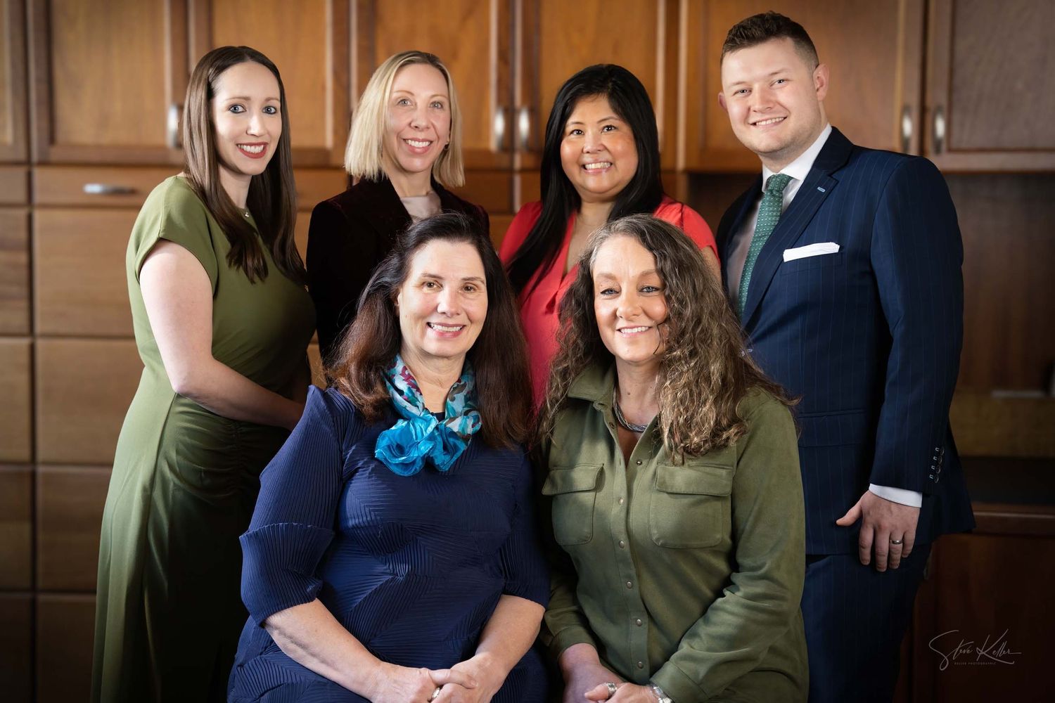 Six professionally dressed colleagues pose for a formal group portrait in an elegant wood-paneled office setting.