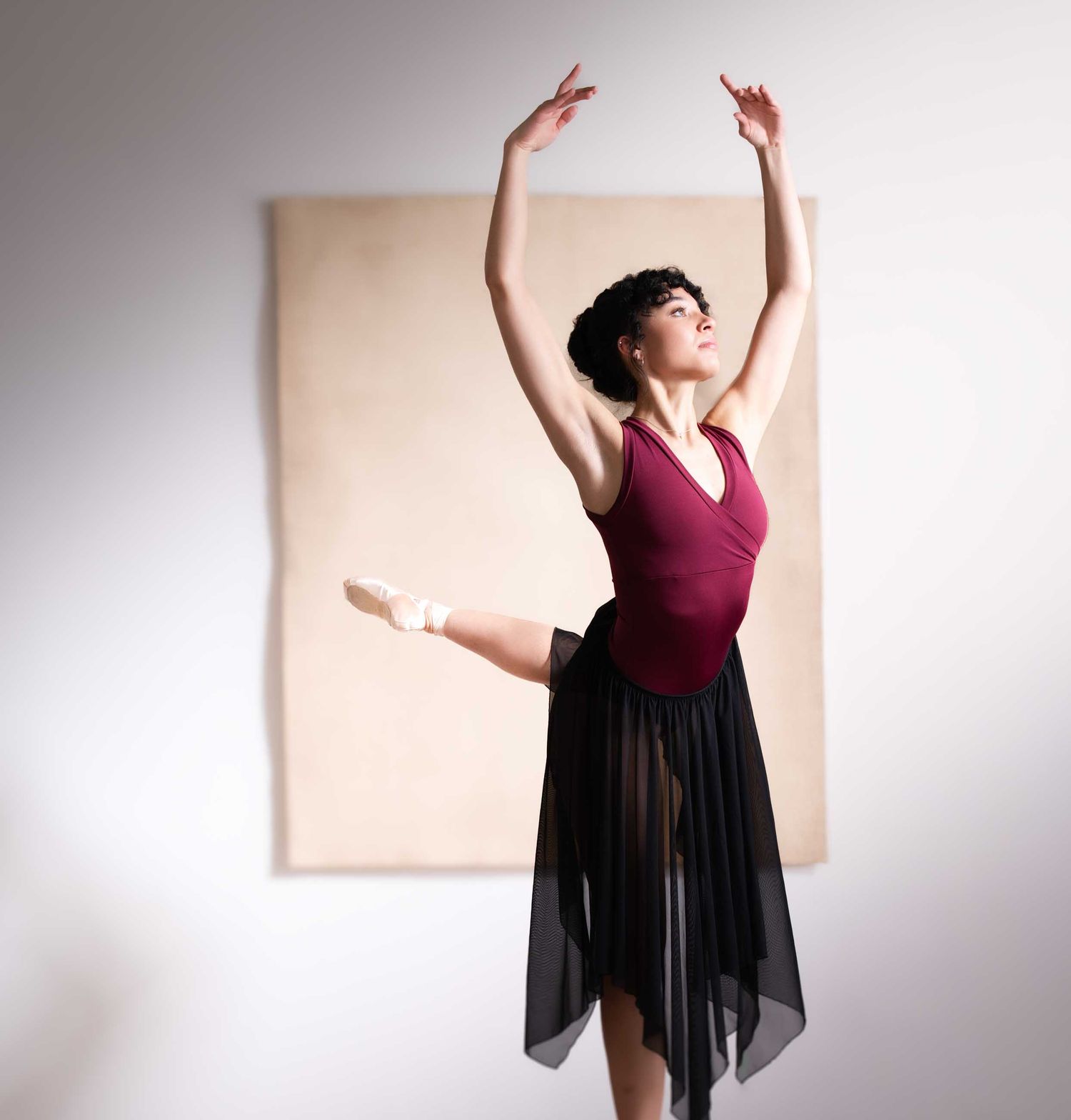 A ballet dancer in a maroon leotard and black skirt performs an arabesque with arms raised gracefully overhead.