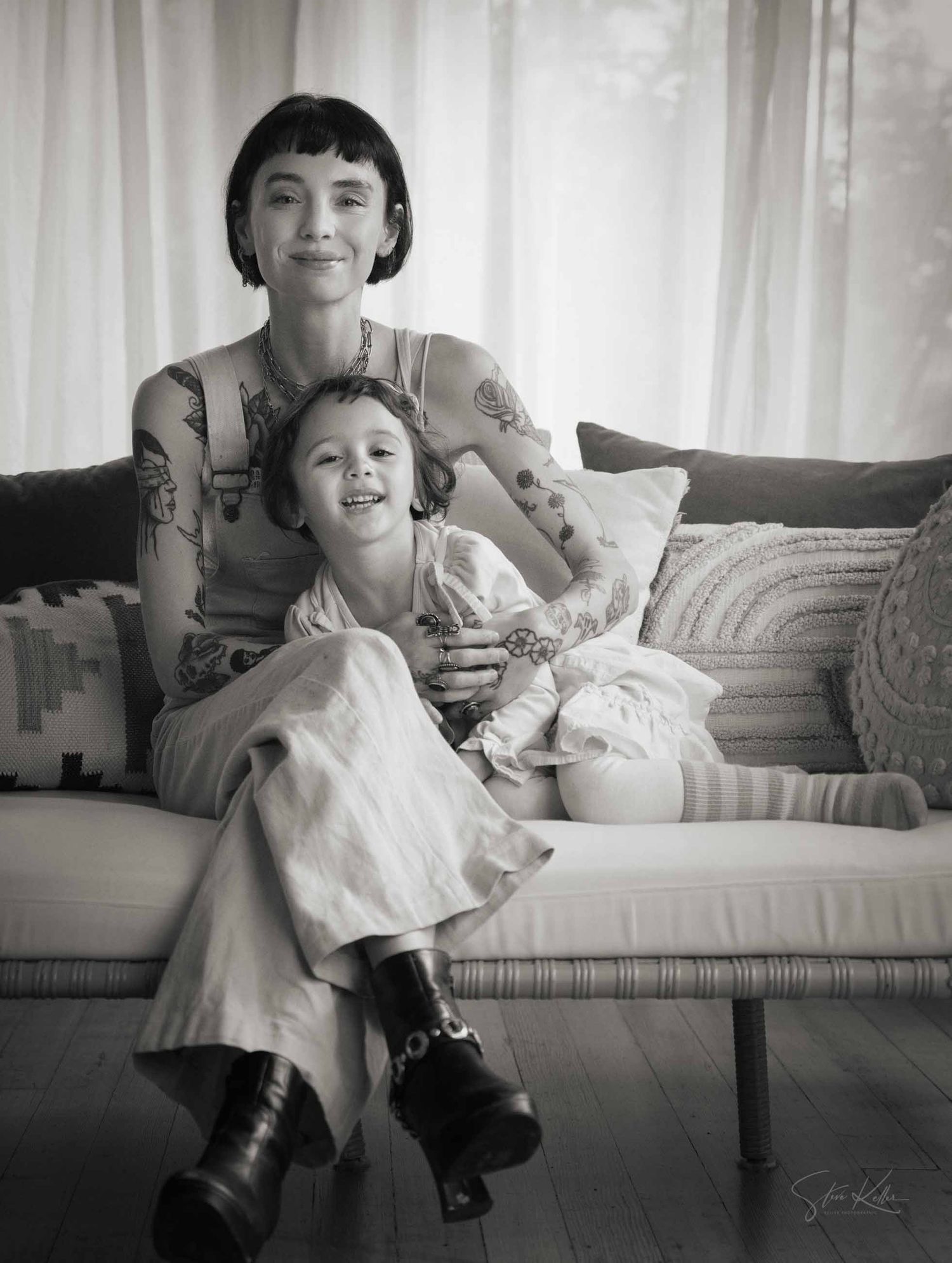 A black and white portrait shows a parent and child relaxing together on a couch in a sunlit room.